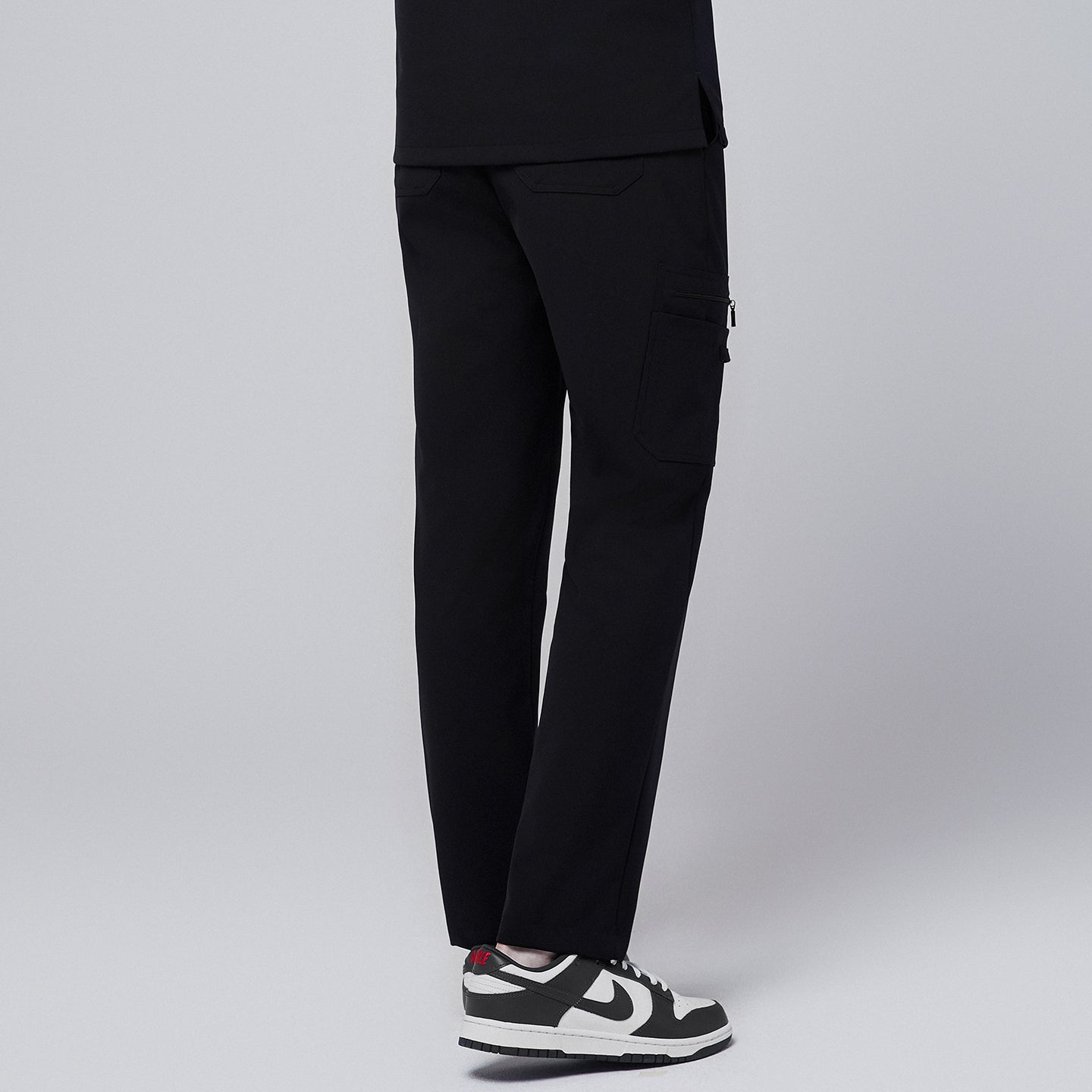 Male model wearing jet black relaxed-fit scrub pants featuring a rear view of the functional side pocket design, paired with black and white sneakers for a professional appearance,Jet Black