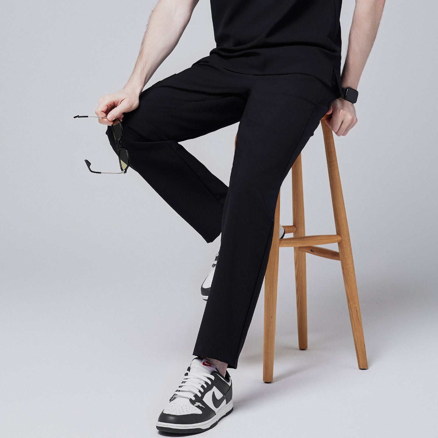 Male model sitting on a wooden stool wearing jet black relaxed-fit scrub pants paired with black and white sneakers, holding a pair of sunglasses for a casual yet professional look,Jet Black