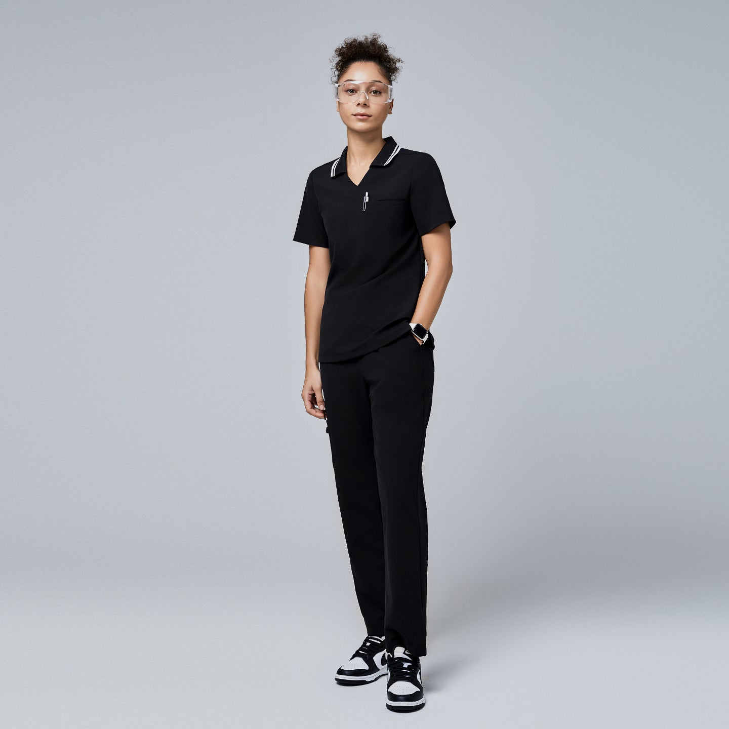 Female model wearing a jet black essential polo scrub top with a striped collar and matching pants, accessorized with safety glasses and sneakers,Jet Black