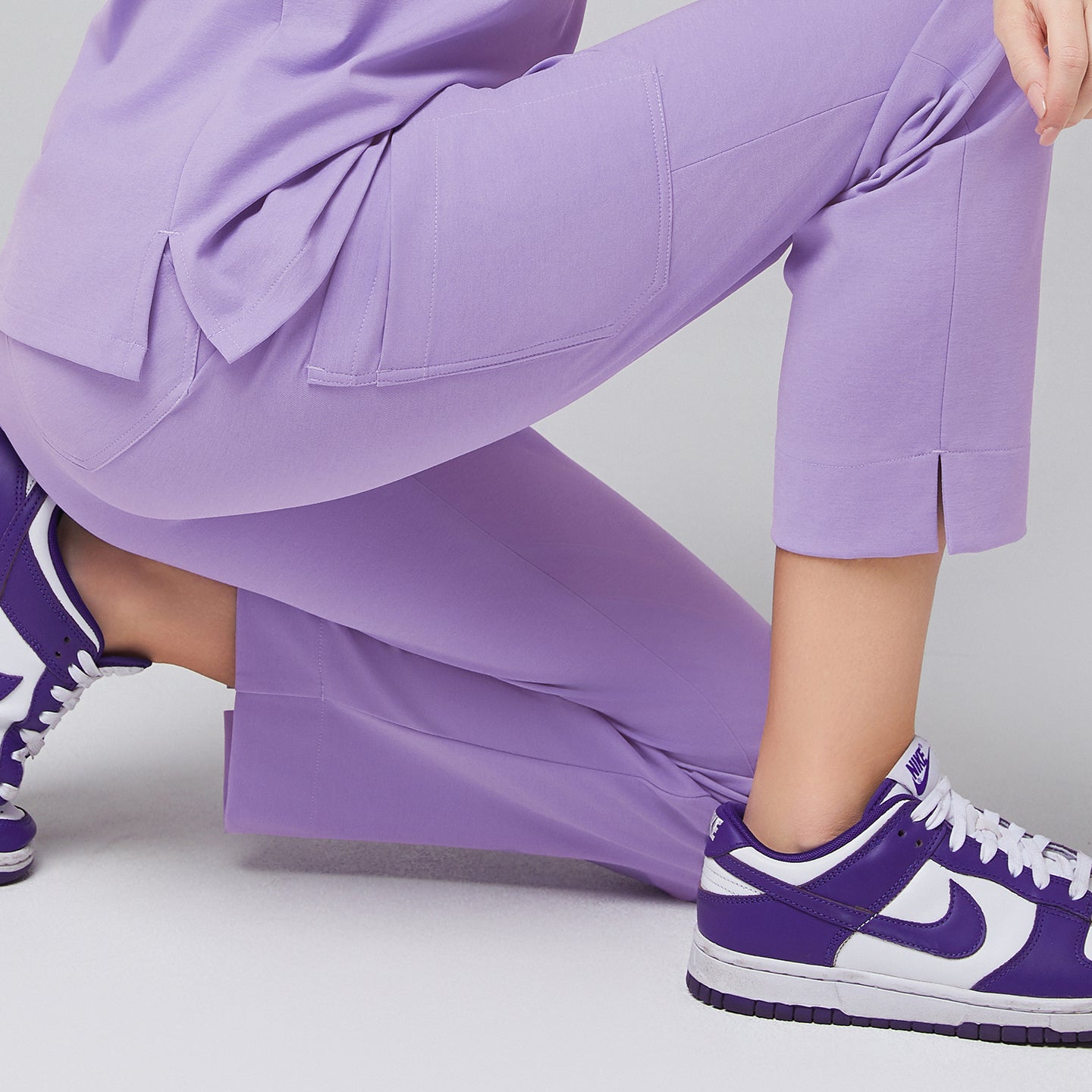 Close-up of lavender scrub pants with split hem detail, shown while kneeling, paired with purple sneakers,Lavender