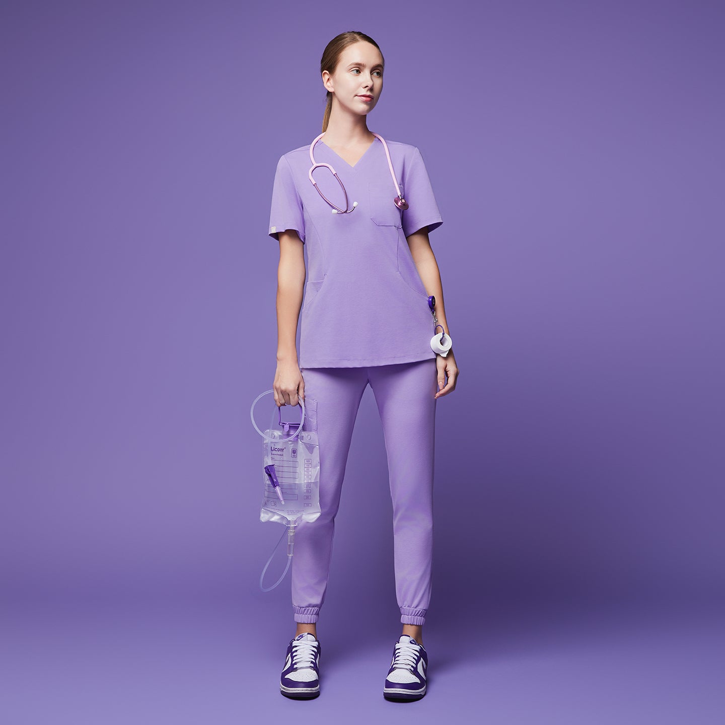 Female wearing lavender jogger scrub pants and top with a stethoscope, holding a medical fluid bag, paired with purple and white sneakers,Lavender