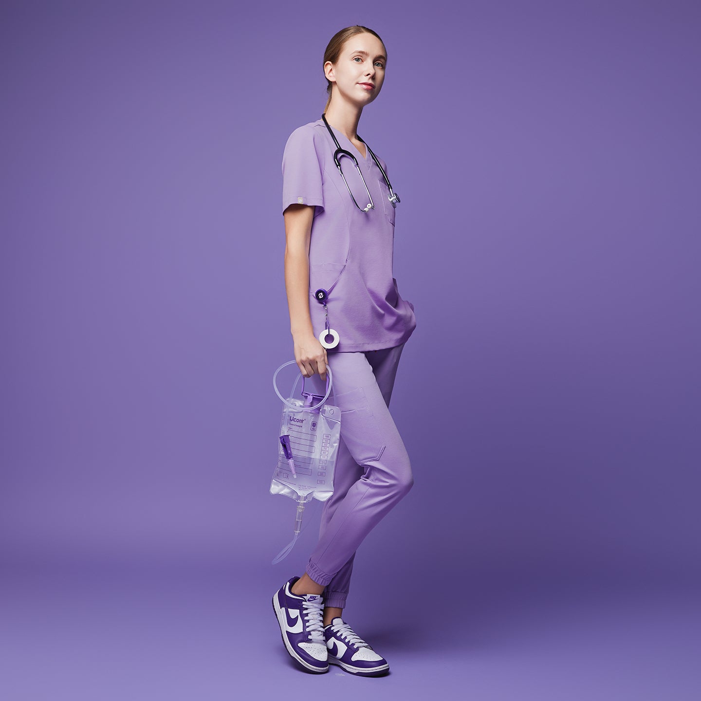 Female wearing lavender jogger scrub pants and top with a stethoscope, holding a medical fluid bag, standing in a side pose, paired with purple and white sneakers,Lavender