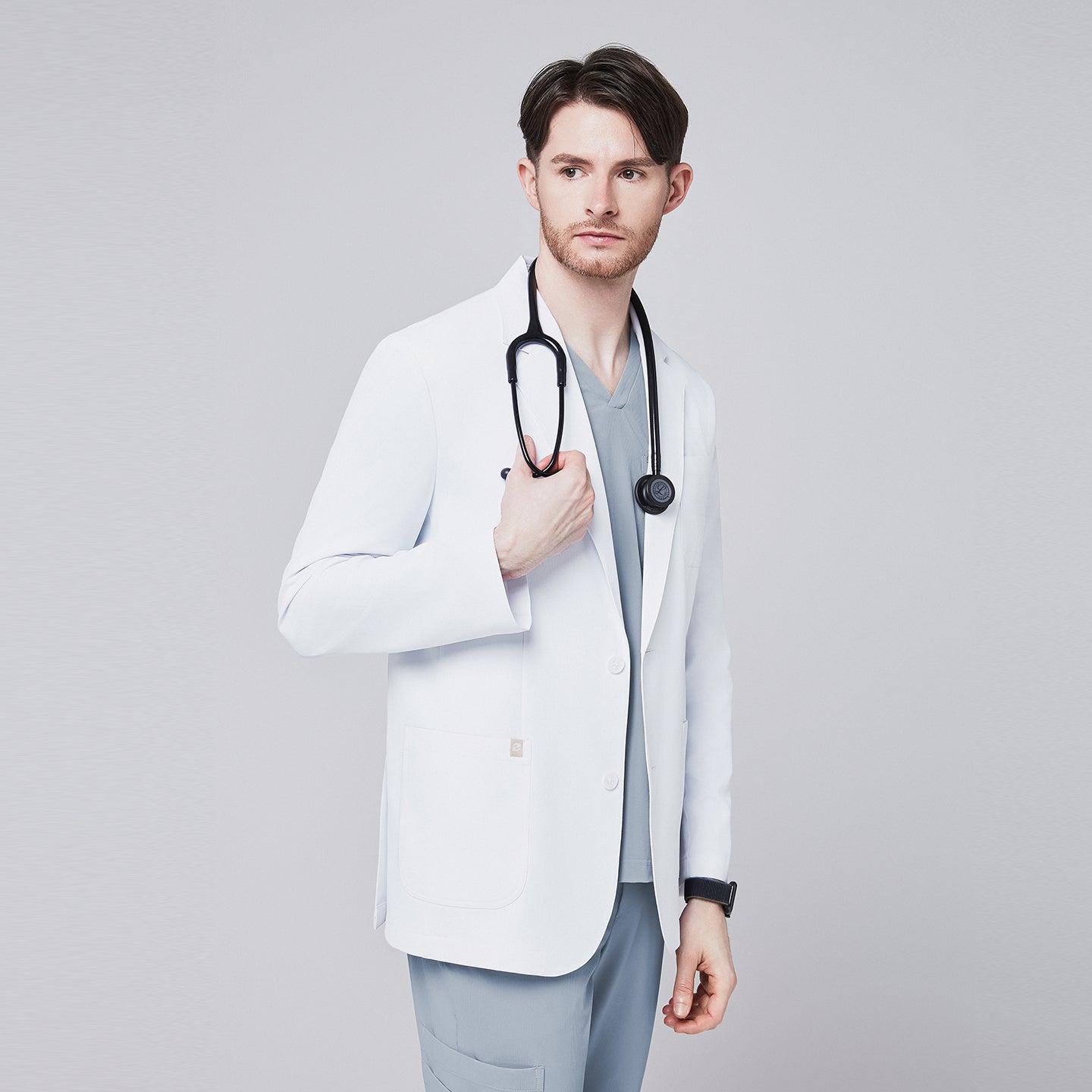 A man wearing a short white lab coat (LCM-01) over a Cool Gray Single Pocket Scrub Top and Cool Gray StretchFit Jogger Scrub Pants, looking to the side while holding a stethoscope,White