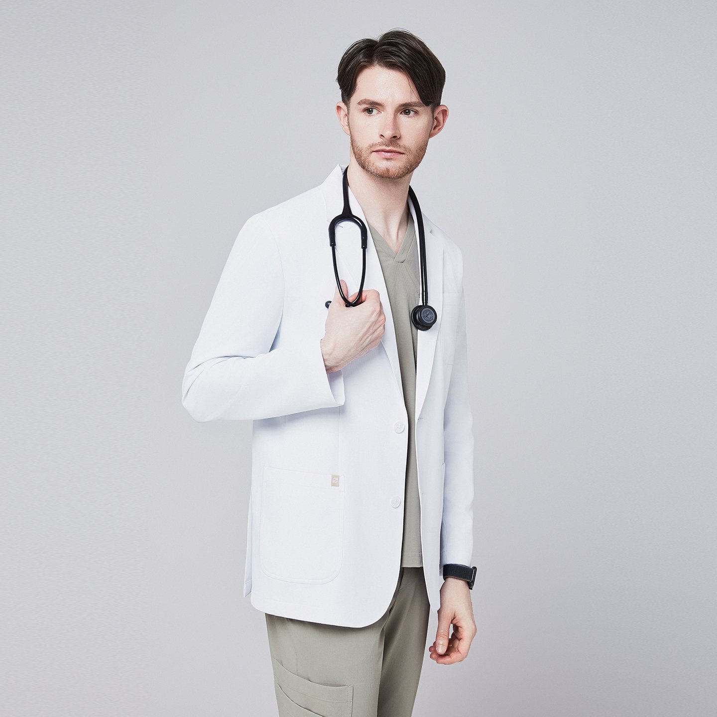 A man wearing a short white lab coat (LCM-01) over a Sand Khaki Single Pocket Scrub Top and Sand Khaki StretchFit Jogger Scrub Pants, holding a stethoscope near his chest,White