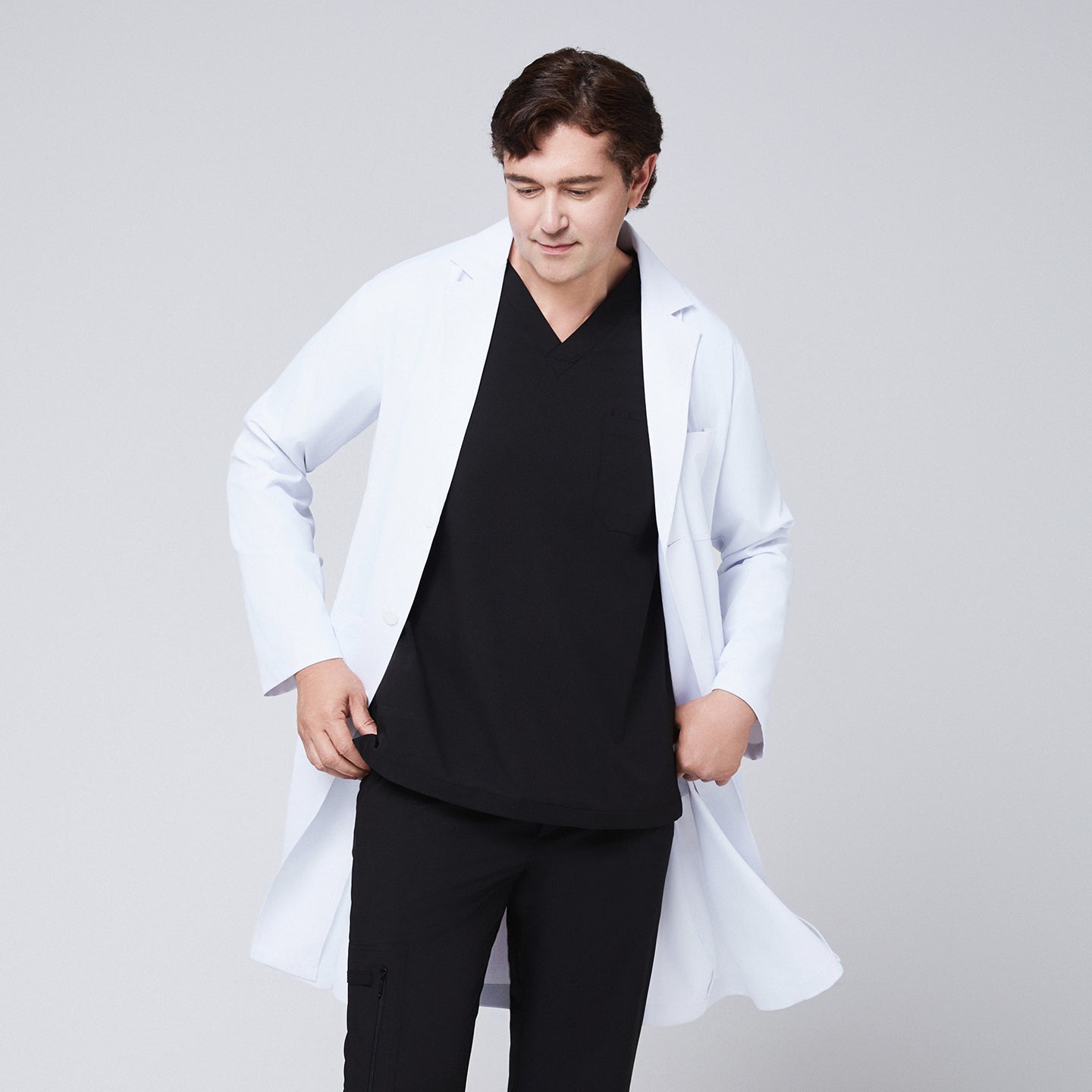 A man wearing a long white lab coat (LCM-02) over an Eco Black Double Pocket Scrub Top and Eco Black Zipper Pocket Straight Scrub Pants, looking down while holding the coat open,White