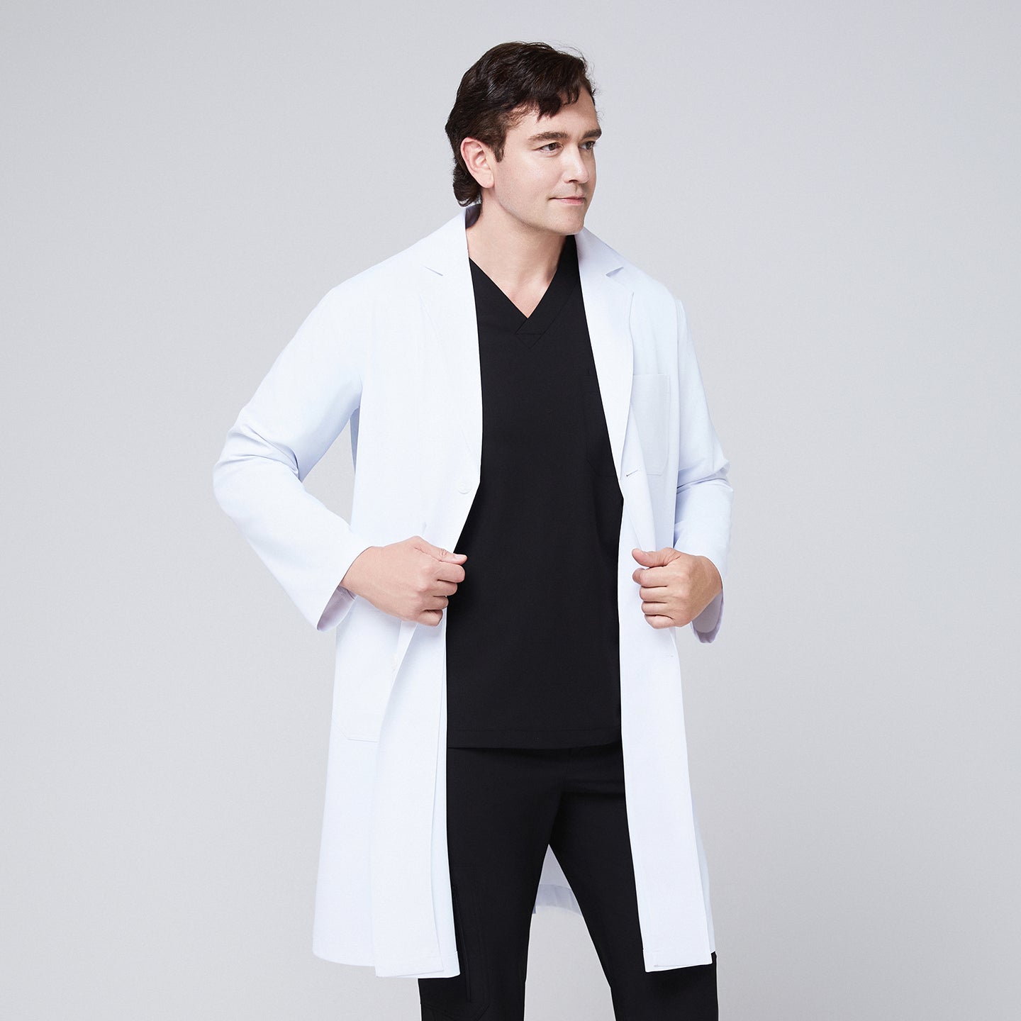 A man wearing a long white lab coat (LCM-02) over an Eco Black Double Pocket Scrub Top and Eco Black Zipper Pocket Straight Scrub Pants, adjusting the front of the coat,White