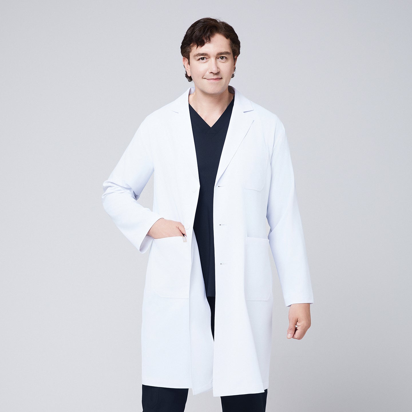 A man wearing a long white lab coat (LCM-02) over an Eco Navy Double Pocket Scrub Top and Eco Navy Zipper Pocket Straight Scrub Pants, placing his hand in the coat pocket,White