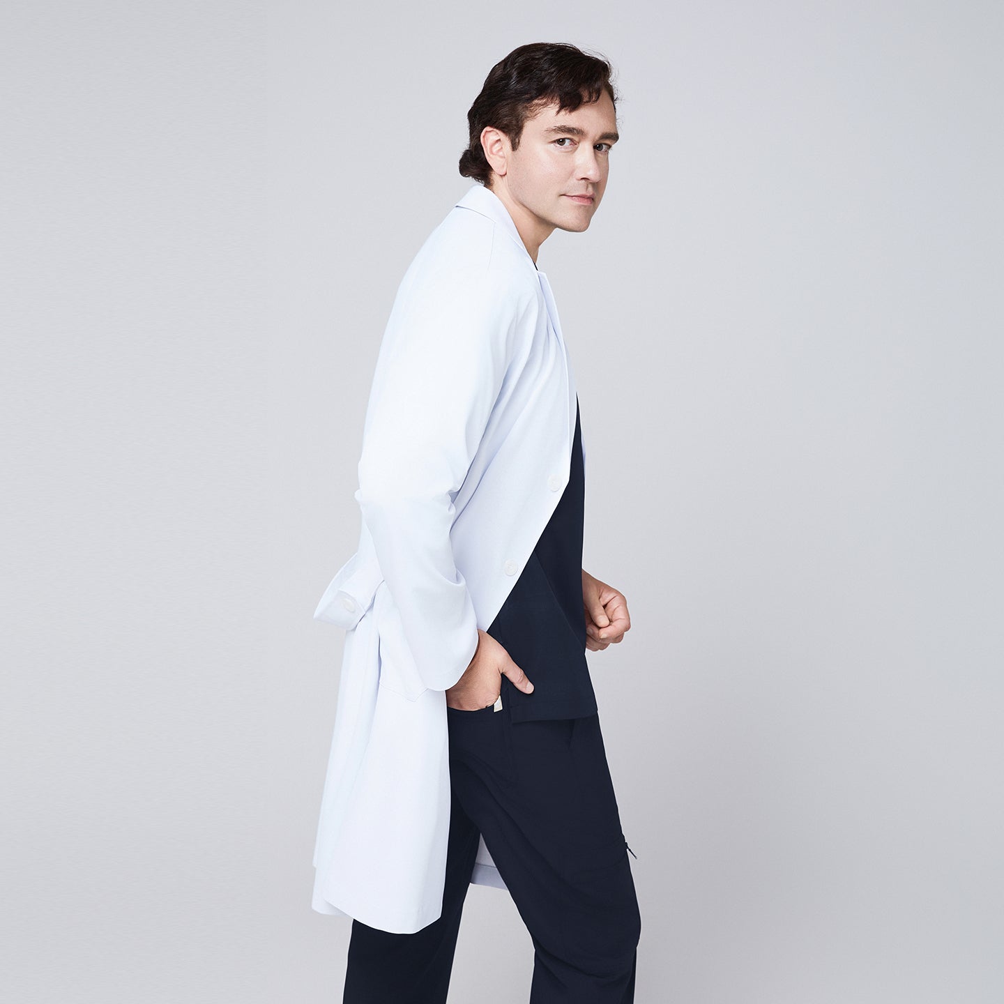 Side view of a man wearing a long white lab coat (LCM-02) over an Eco Navy Double Pocket Scrub Top and Eco Navy Zipper Pocket Straight Scrub Pants, looking toward the camera,Eco Navy