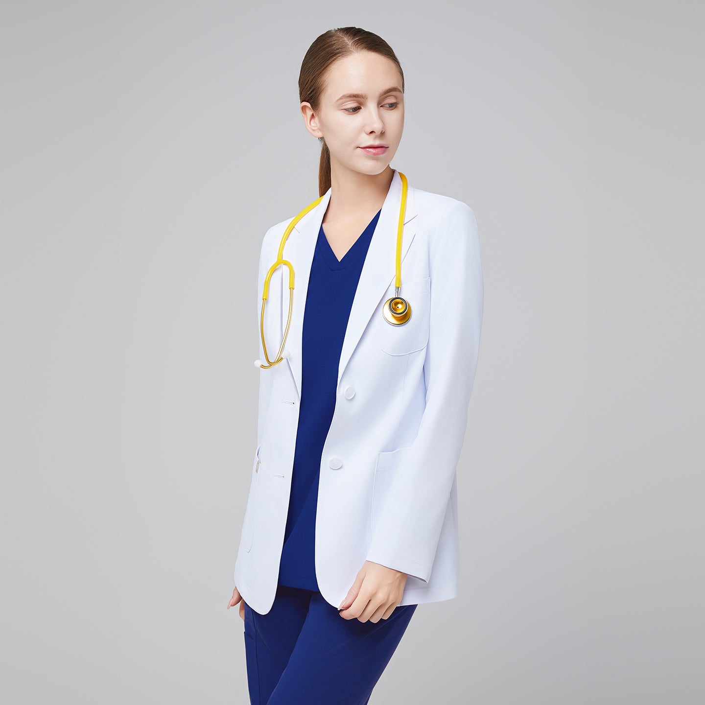 A woman wearing a short white lab coat (LCW-02) over an Admiral Blue Double Pocket Scrub Top and Admiral Blue StretchFit Jogger Scrub Pants, looking to the side with a yellow stethoscope around her neck,White