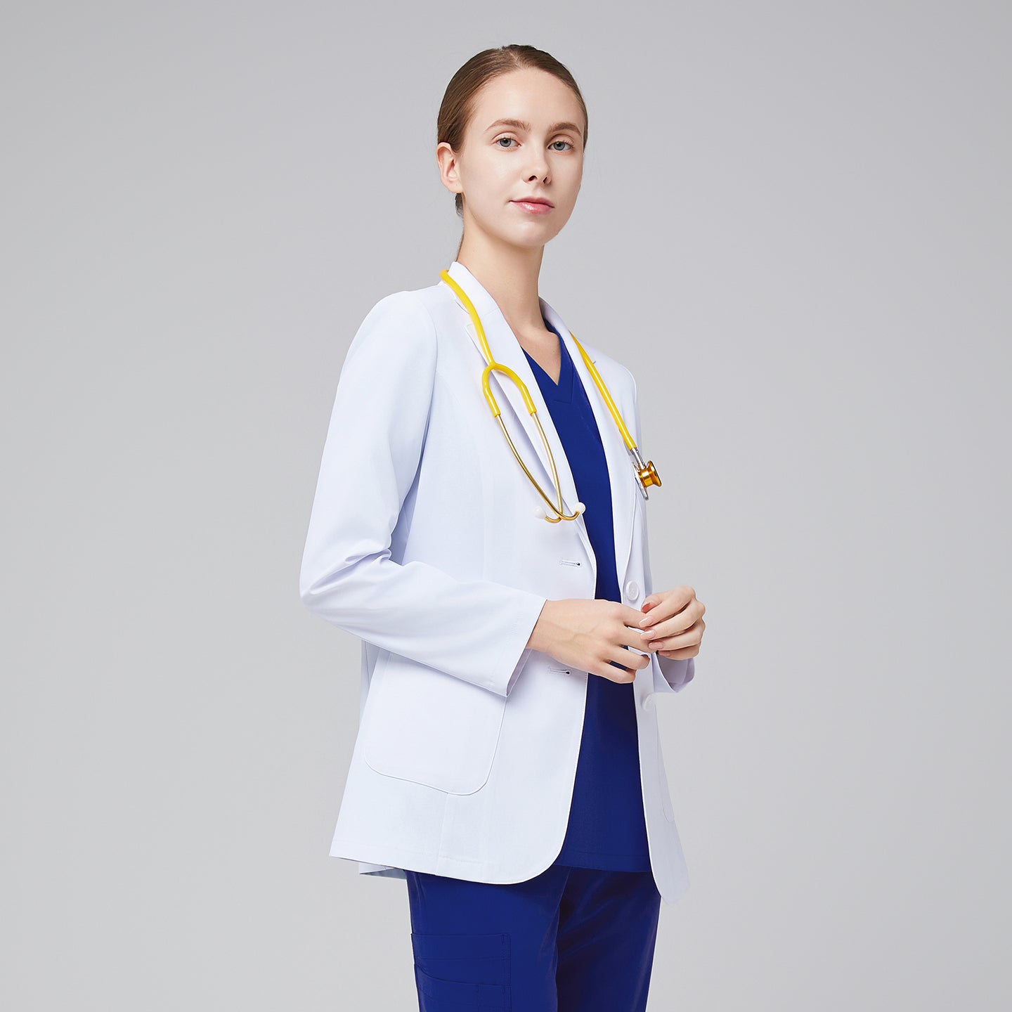 A woman wearing a short white lab coat (LCW-02) over an Admiral Blue Double Pocket Scrub Top and Admiral Blue StretchFit Jogger Scrub Pants, looking straight ahead while holding a yellow stethoscope,White