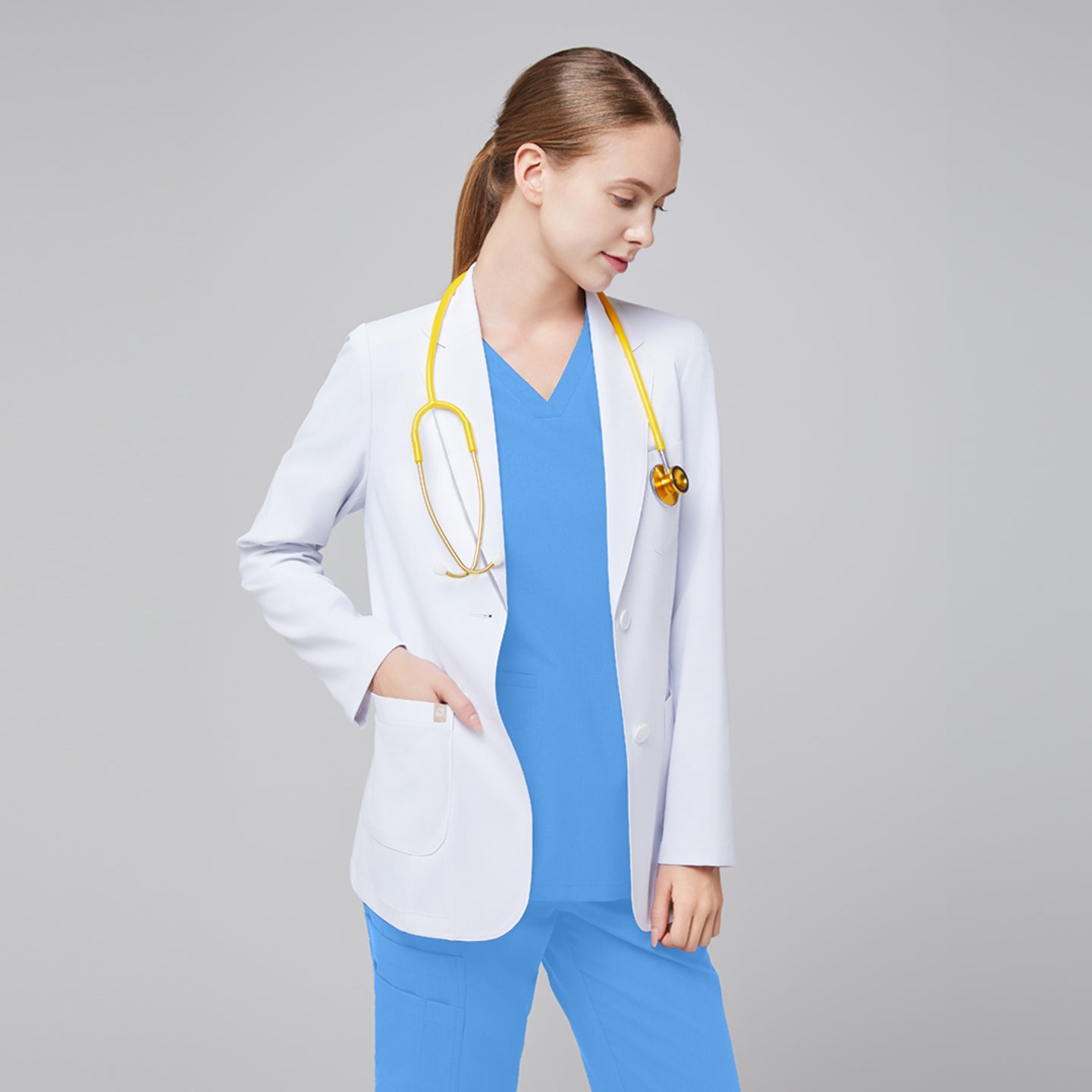 A woman wearing a short white lab coat (LCW-02) over a Ceil Blue Double Pocket Scrub Top and Ceil Blue Zipper Pocket Jogger Scrub Pants, with a yellow stethoscope around her neck,White