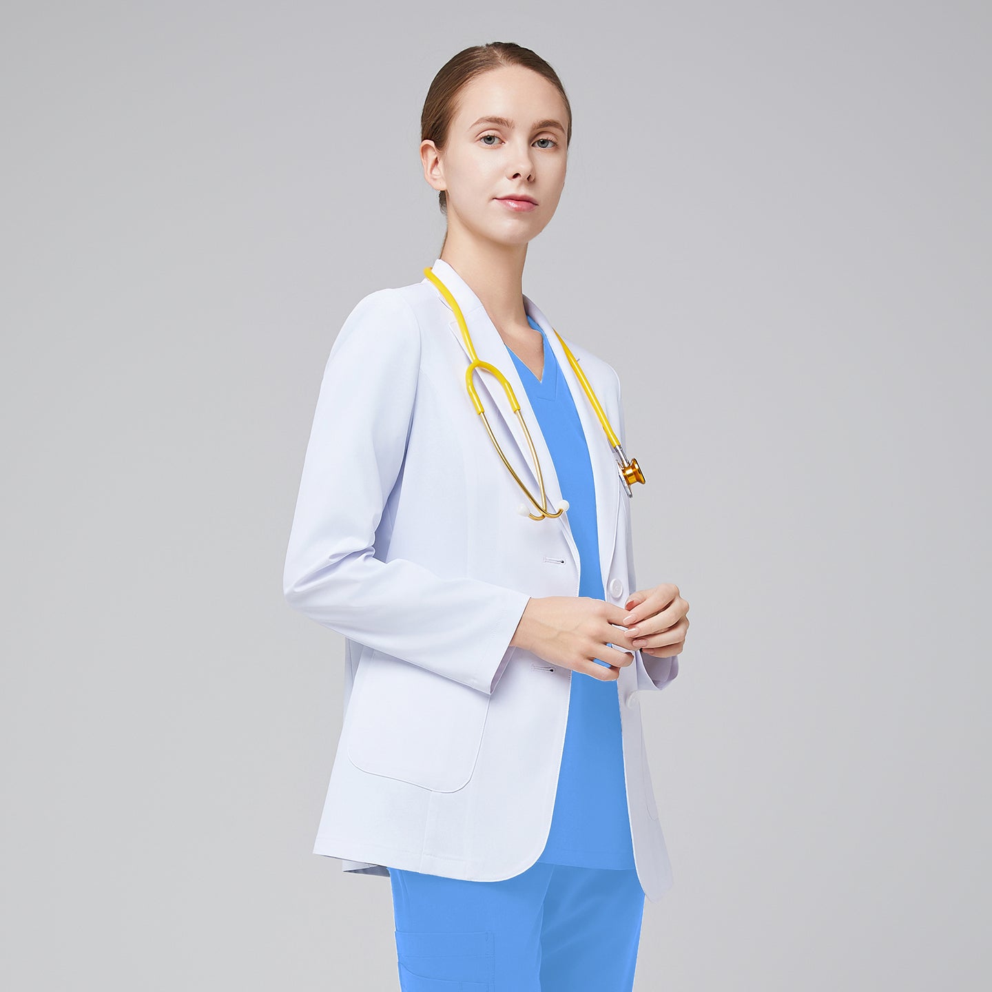 A woman wearing a short white lab coat (LCW-02) over a Ceil Blue Double Pocket Scrub Top and Ceil Blue Zipper Pocket Jogger Scrub Pants, looking straight ahead while holding a yellow stethoscope,White