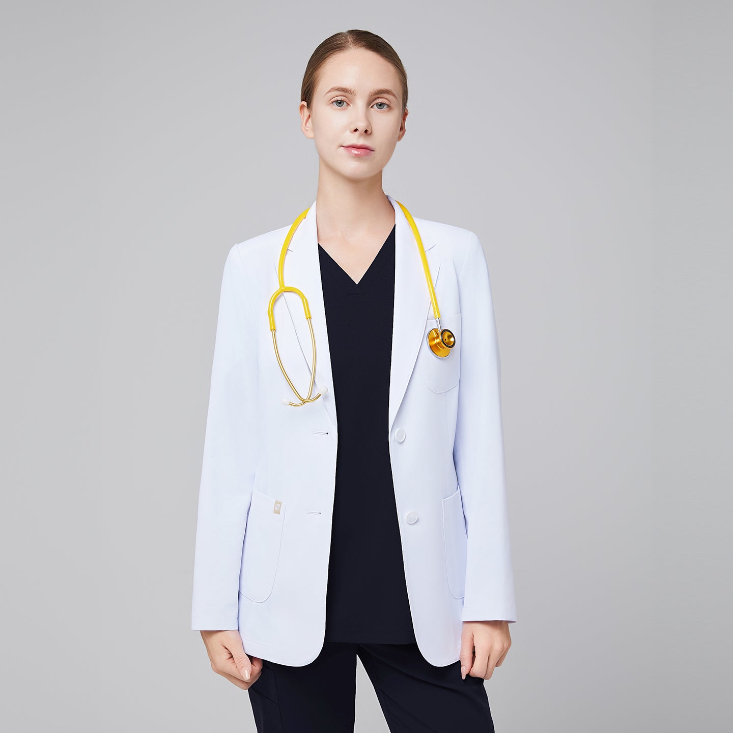 A woman wearing a short white lab coat (LCW-02) over an Eco Navy Double Pocket Scrub Top and Eco Navy Zipper Pocket Jogger Scrub Pants, with a stethoscope around her neck,White