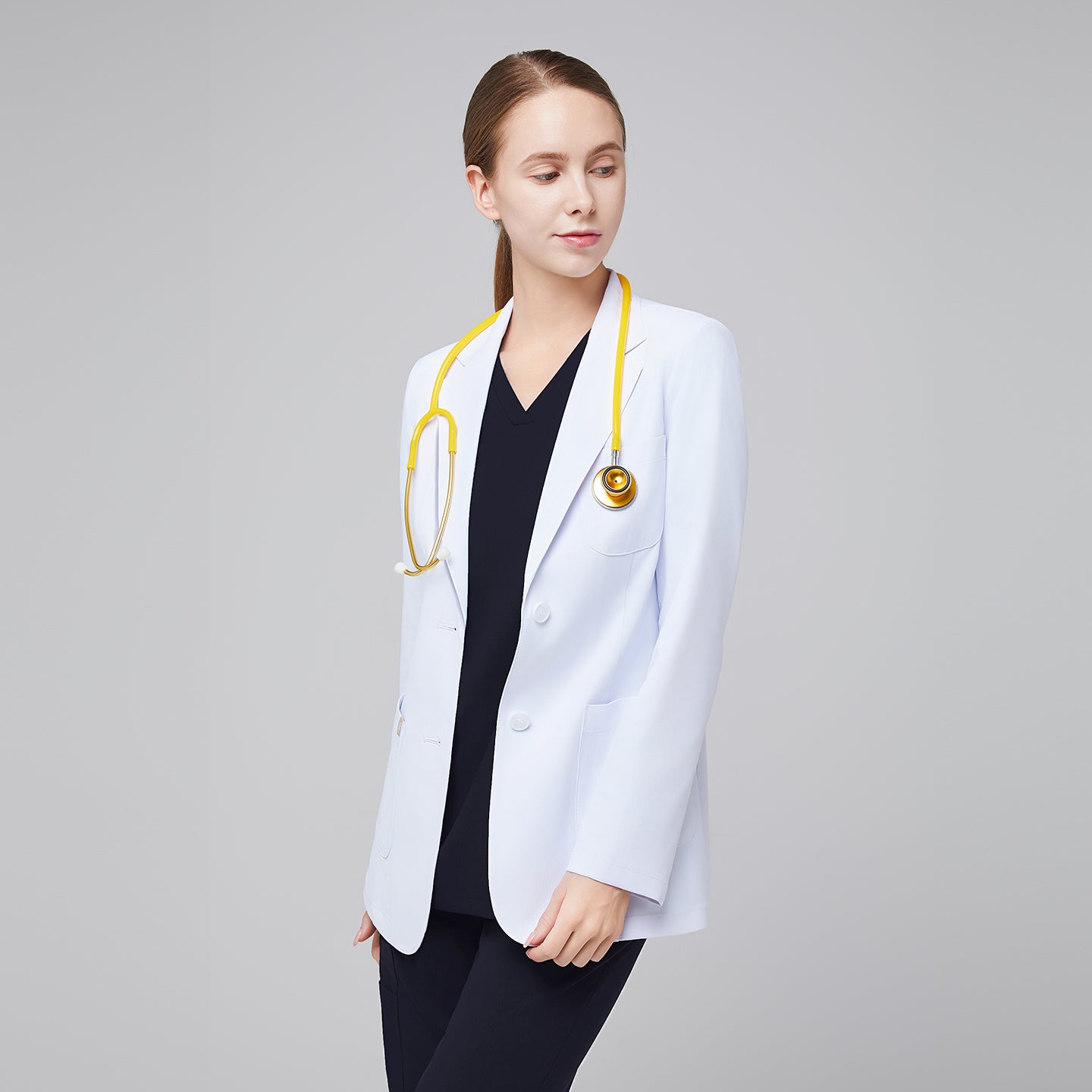 A woman wearing a short white lab coat (LCW-02) over an Eco Navy Double Pocket Scrub Top and Eco Navy Zipper Pocket Jogger Scrub Pants, looking to the side while holding a stethoscope,White