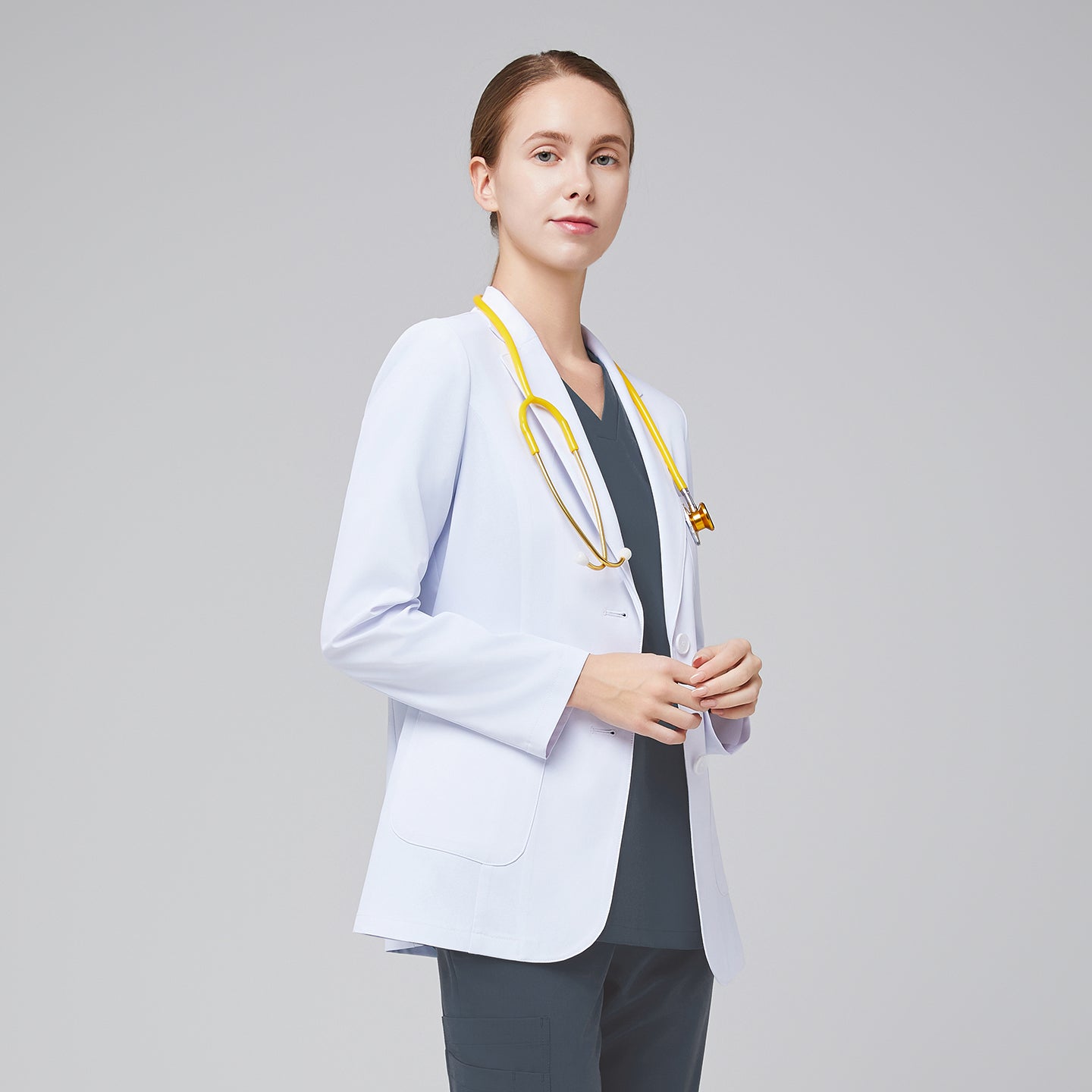 A woman wearing a short white lab coat (LCW-02) over a Smoky Blue Double Pocket Scrub Top and Smoky Blue Straight Split Hem Scrub Pants, looking straight ahead while holding a yellow stethoscope,White