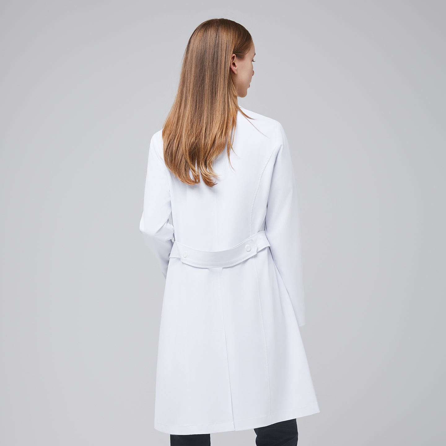 Back view of a woman wearing a long white lab coat (LCW-04), highlighting the buttoned belt detail, over Deep Gray scrubs,White