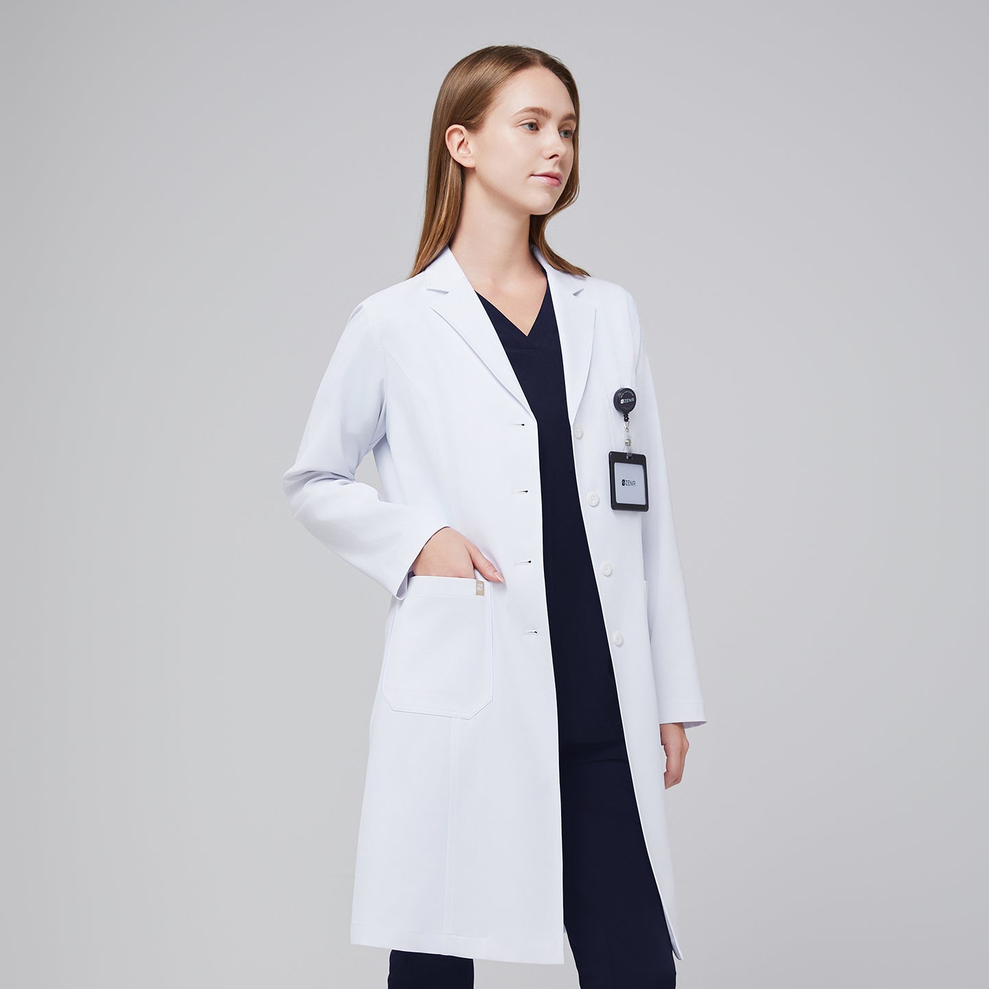 A woman wearing a long white lab coat (LCW-04) over an Eco Navy Single Pocket Scrub Top and Eco Navy Zipper Pocket Jogger Scrub Pants, with an ID badge attached to the coat,White