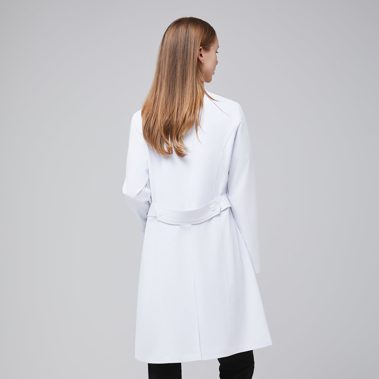 Back view of a woman wearing a long white lab coat (LCW-04) with a buttoned back belt detail, over Eco Navy scrubs,White