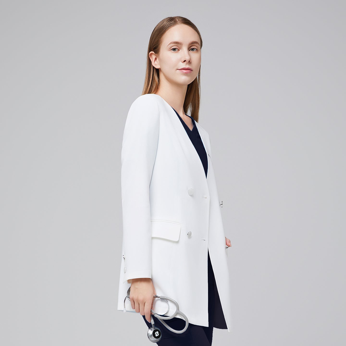 Side view of a woman wearing a white long lab coat (LCW-09) over an Eco Navy Pleated Shoulder Scrub Top and Eco Navy Zipper Pocket Jogger Scrub Pants, holding a stethoscope,White