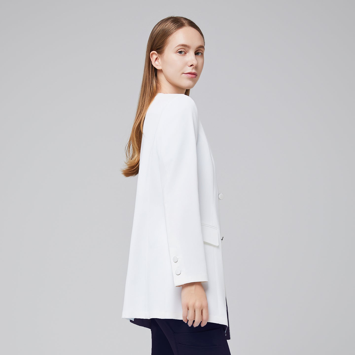 Side view of a woman wearing a white long lab coat (LCW-09) over an Eco Navy Pleated Shoulder Scrub Top and Eco Navy Zipper Pocket Jogger Scrub Pants,White