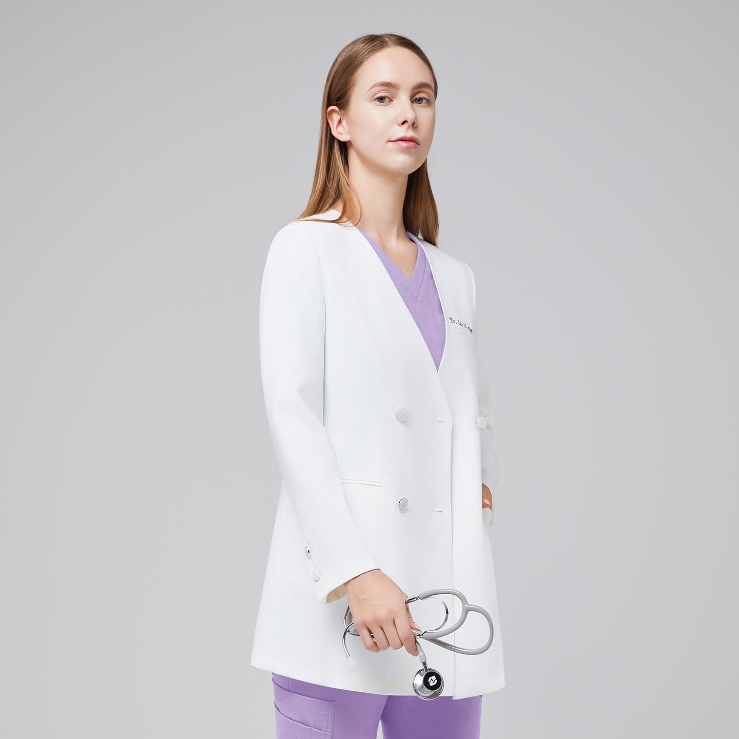 A woman wearing a white long lab coat (LCW-09) over a Lavender Pleated Shoulder Scrub Top and Lavender Straight Split Hem Scrub Pants, holding a stethoscope,White