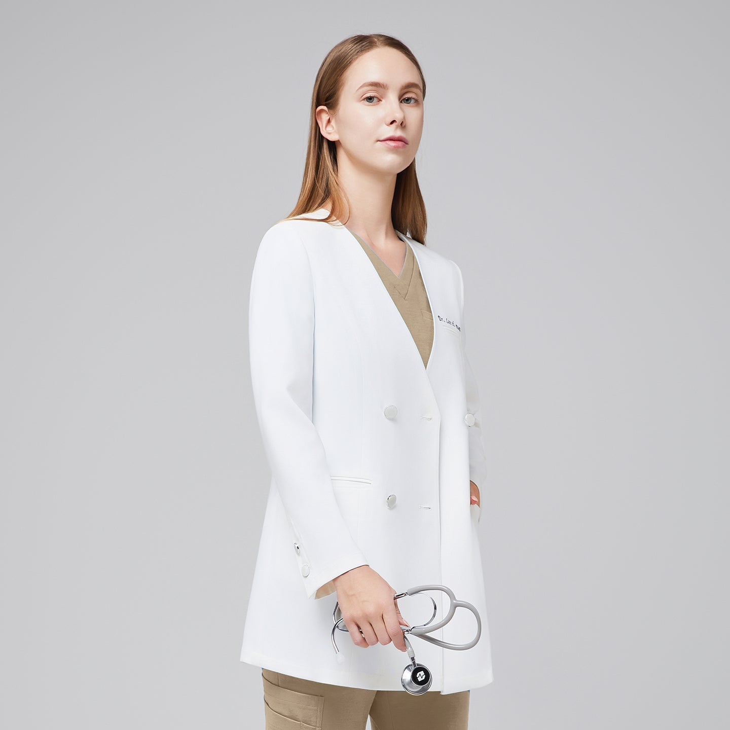 A woman wearing a white long lab coat (LCW-09) over a Sand Beige Pleated Shoulder Scrub Top and Sand Beige Straight Split Hem Scrub Pants, holding a stethoscope in her hand,White