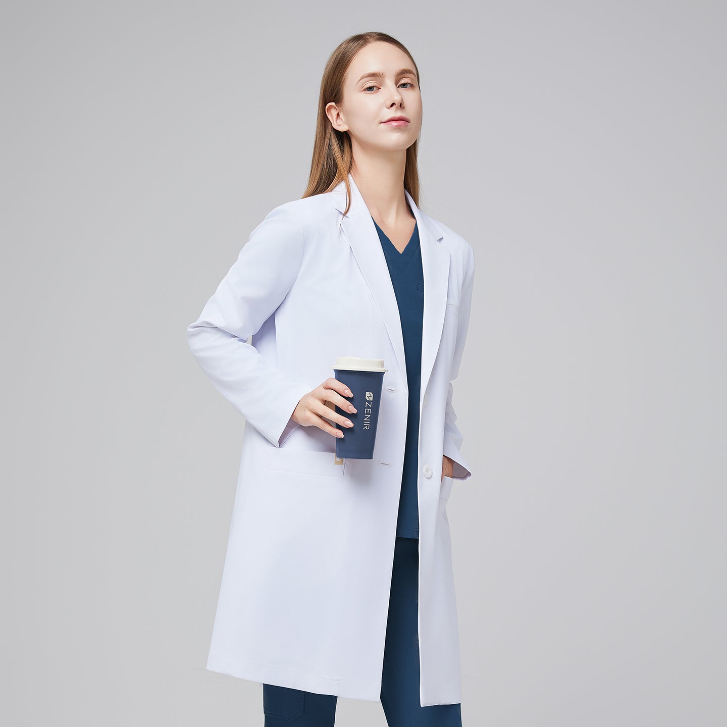 A woman wearing a white long lab coat (LCW-10) over a Dark Blue Soft Stretch Scrub Top and Dark Blue Split Hem Scrub Pants, holding a coffee cup,White












