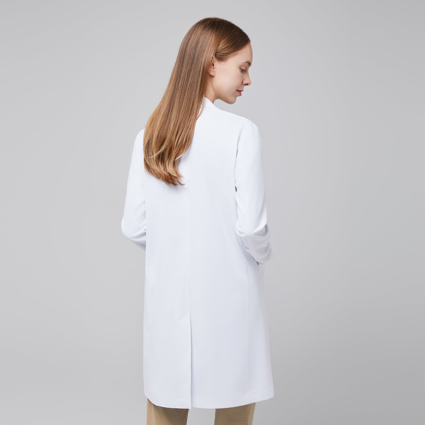 Back view of a woman wearing Zenir’s Women’s White Long Lab Coat LCW-10 over Sand Beige scrubs, highlighting the coat’s length and back design,White