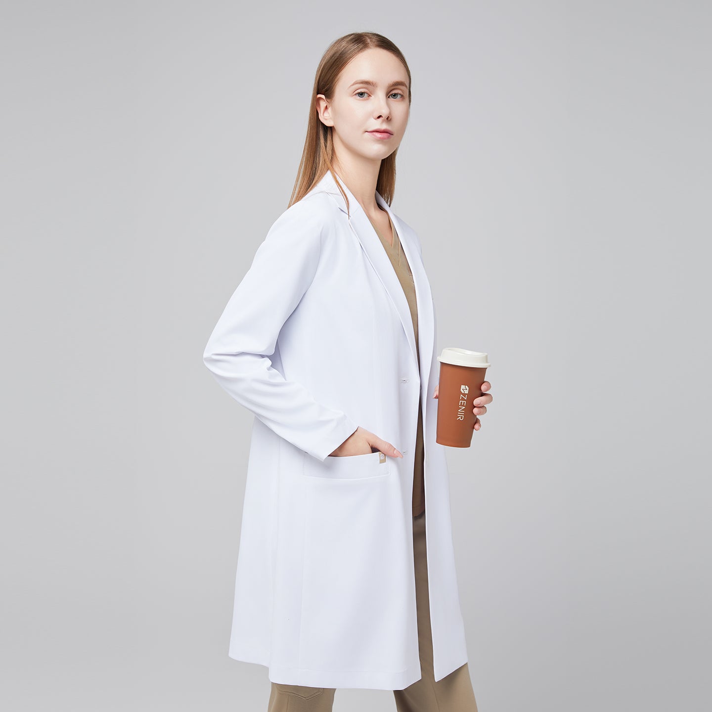 A woman wearing Zenir’s Women’s White Long Lab Coat LCW-10 over a Sand Beige Scrub Top and Pants, holding a coffee cup,White