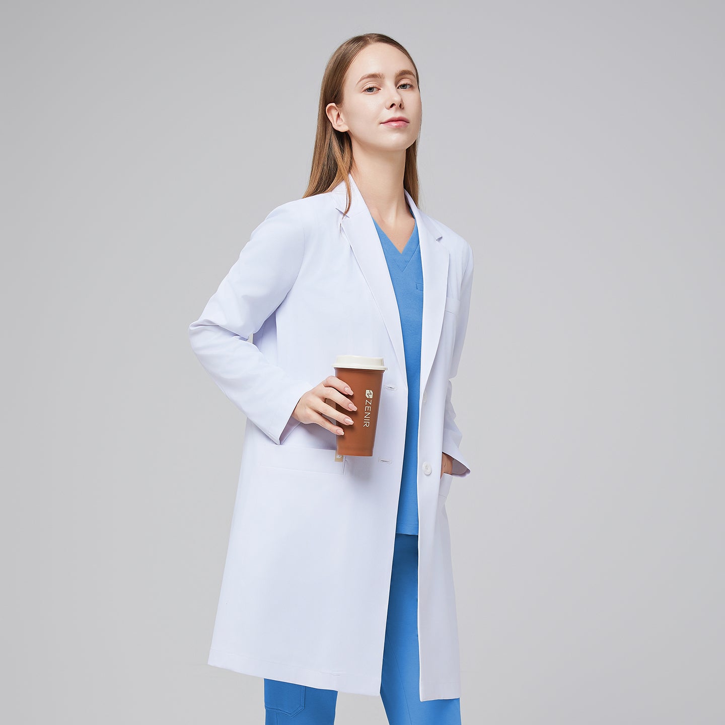 A woman wearing a white women’s Long Lab Coat LCW-10 over a Sky Blue Soft Stretch Scrub Top and Sky Blue Split Hem Scrub Pants, holding a coffee cup,White