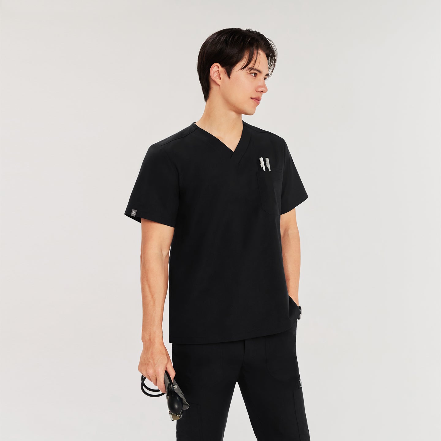 Man in black Zenir scrub top with chest pocket holding pens, standing with  sphygmomanometer in hand, looking to the side,Black