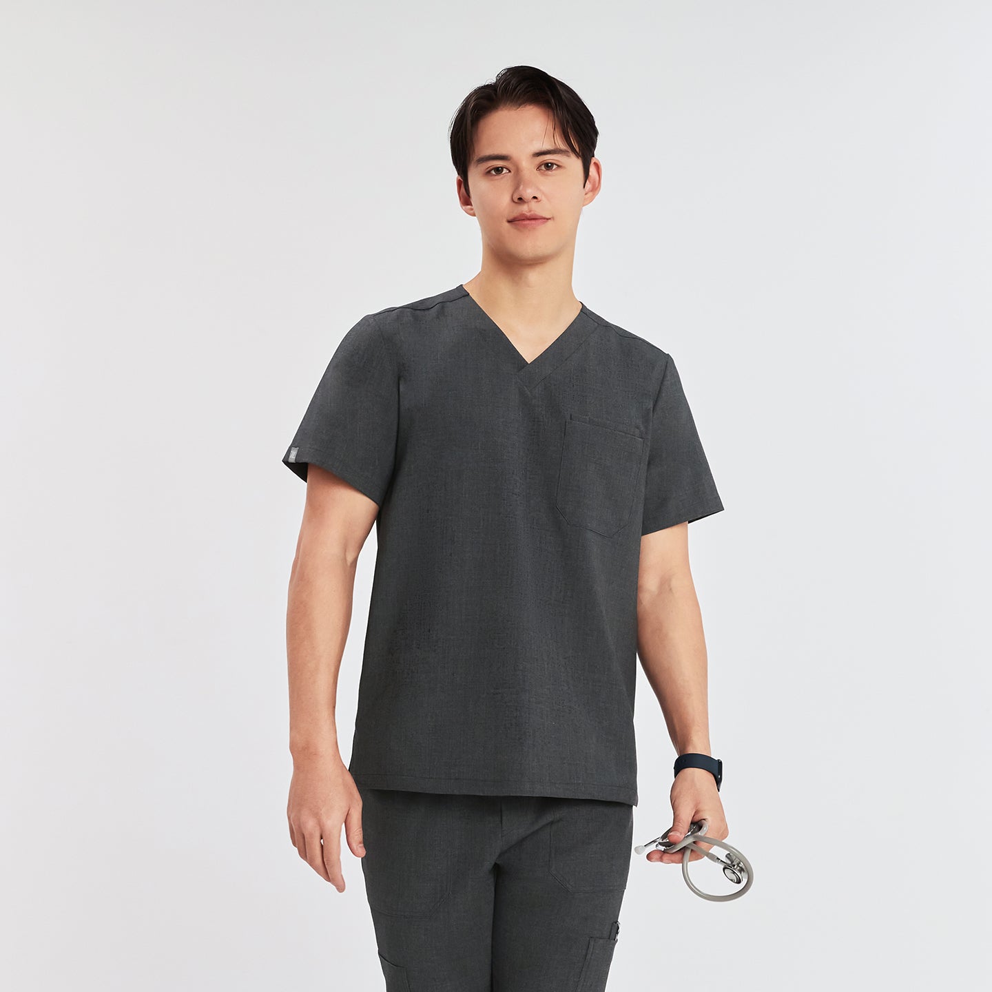 A man wearing a 3-pocket scrub top, standing and holding a stethoscope in one hand,Charcoal Gray
