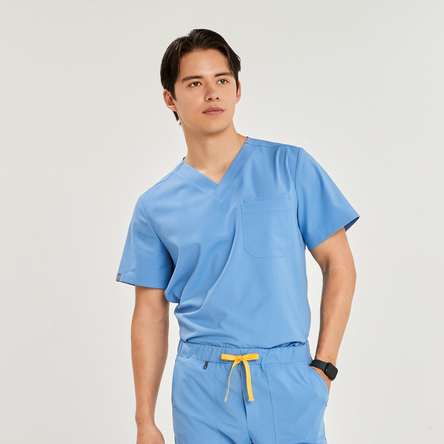 A man wearing a V-neck scrub top with a front chest pocket, standing with one hand in his pocket,Ceil Blue