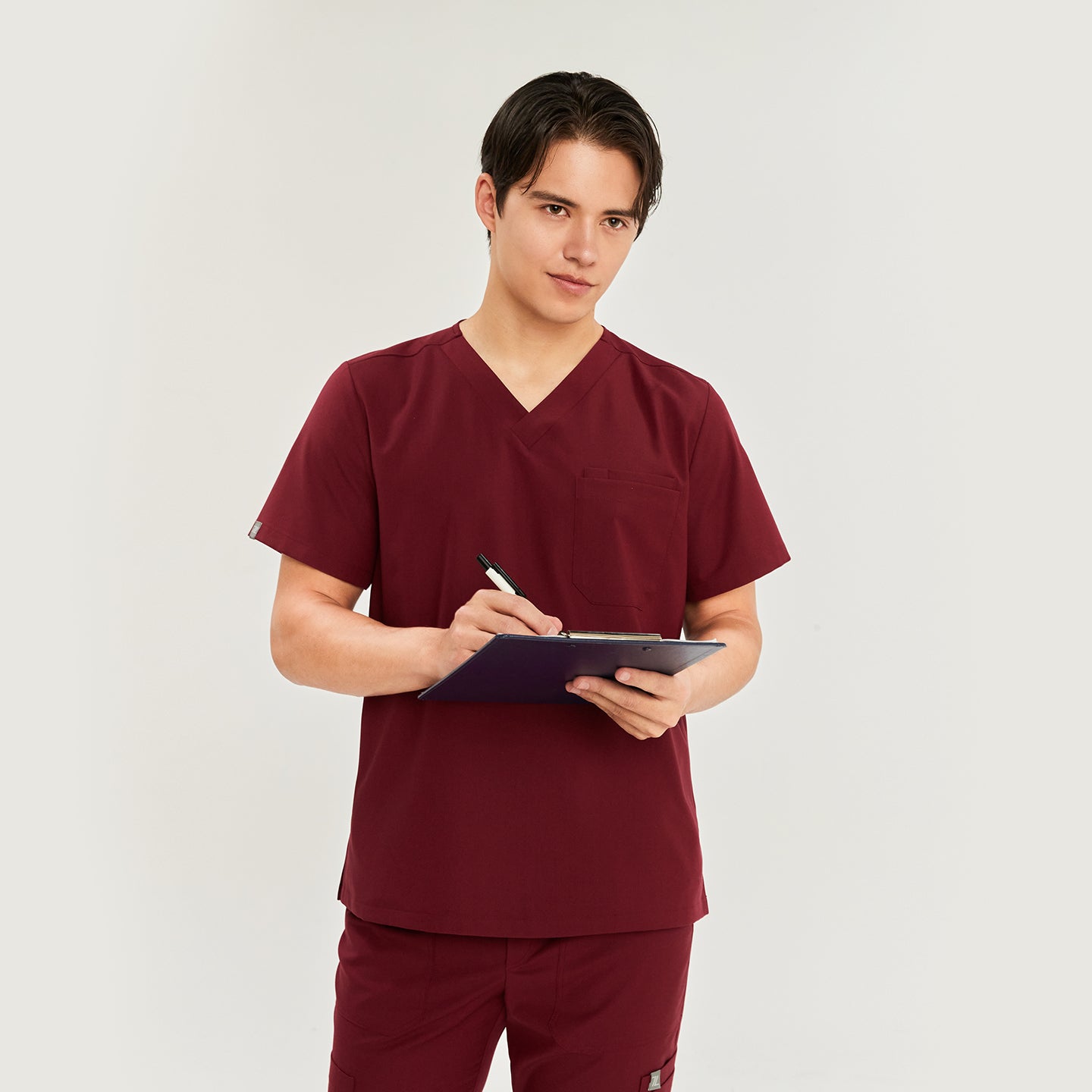 Male model wearing a scrub top with a chest pocket, paired with matching scrub pants with a drawstring, looking to the side,Burgundy