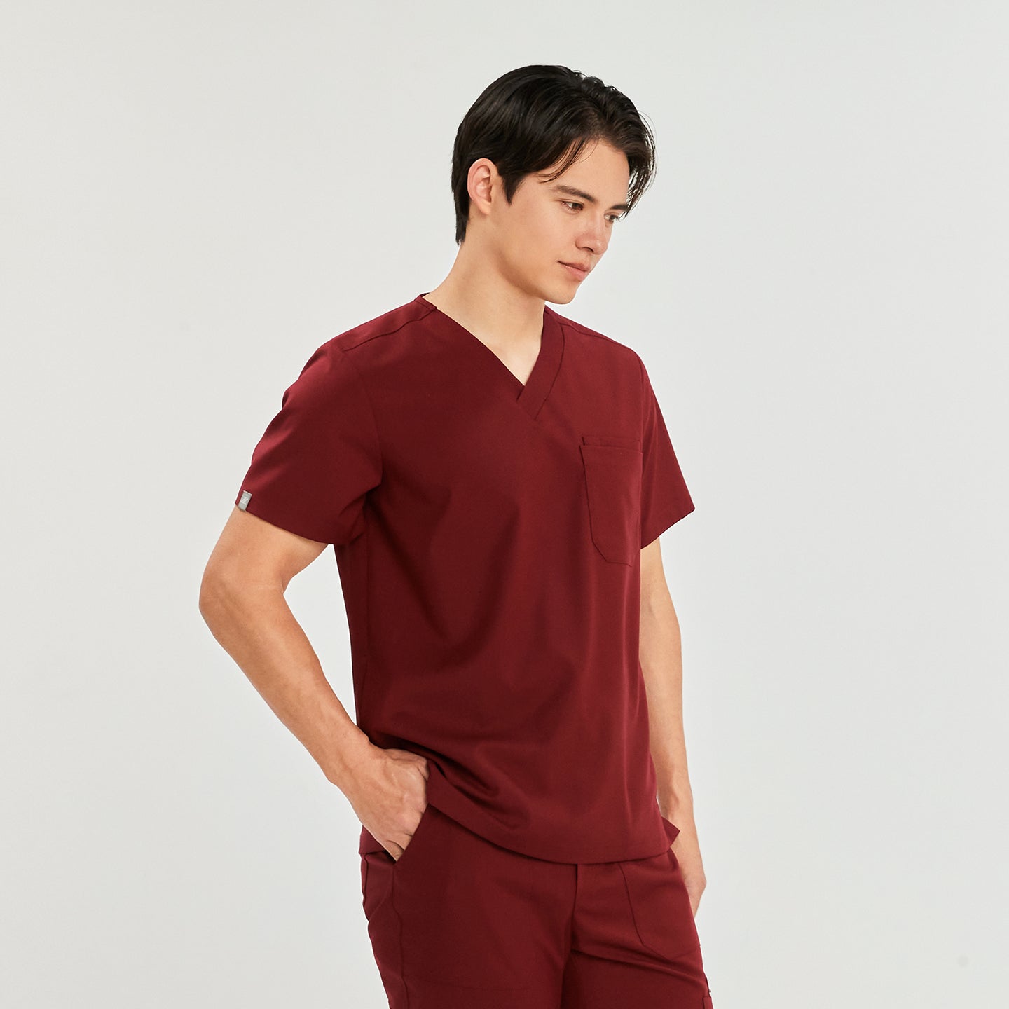  A man wearing a V-neck scrub top with a front chest pocket, standing with his hands in his pockets,Burgundy
