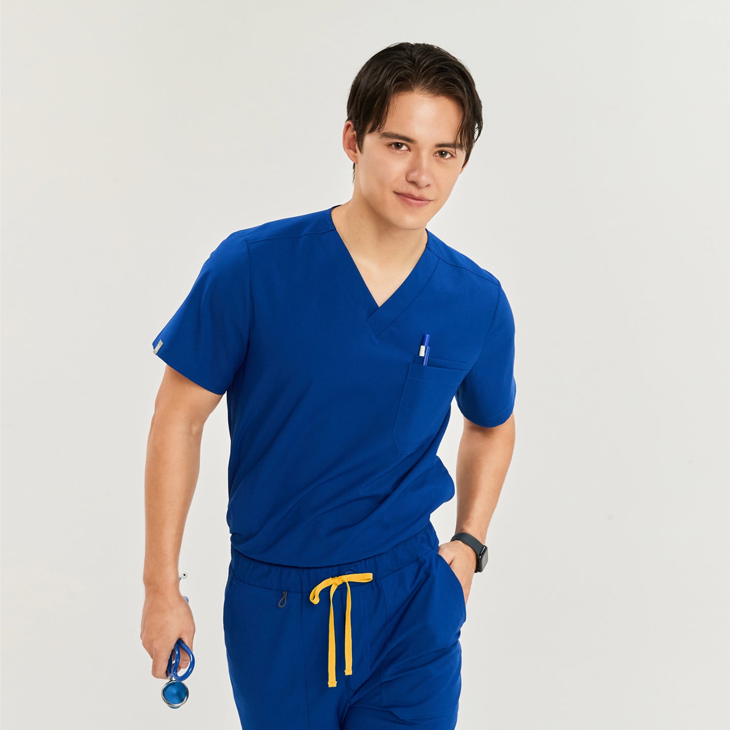 Male model wearing a scrub top with a chest pocket and drawstring pants, holding a stethoscope, and smiling while looking at the camera, Royal Blue