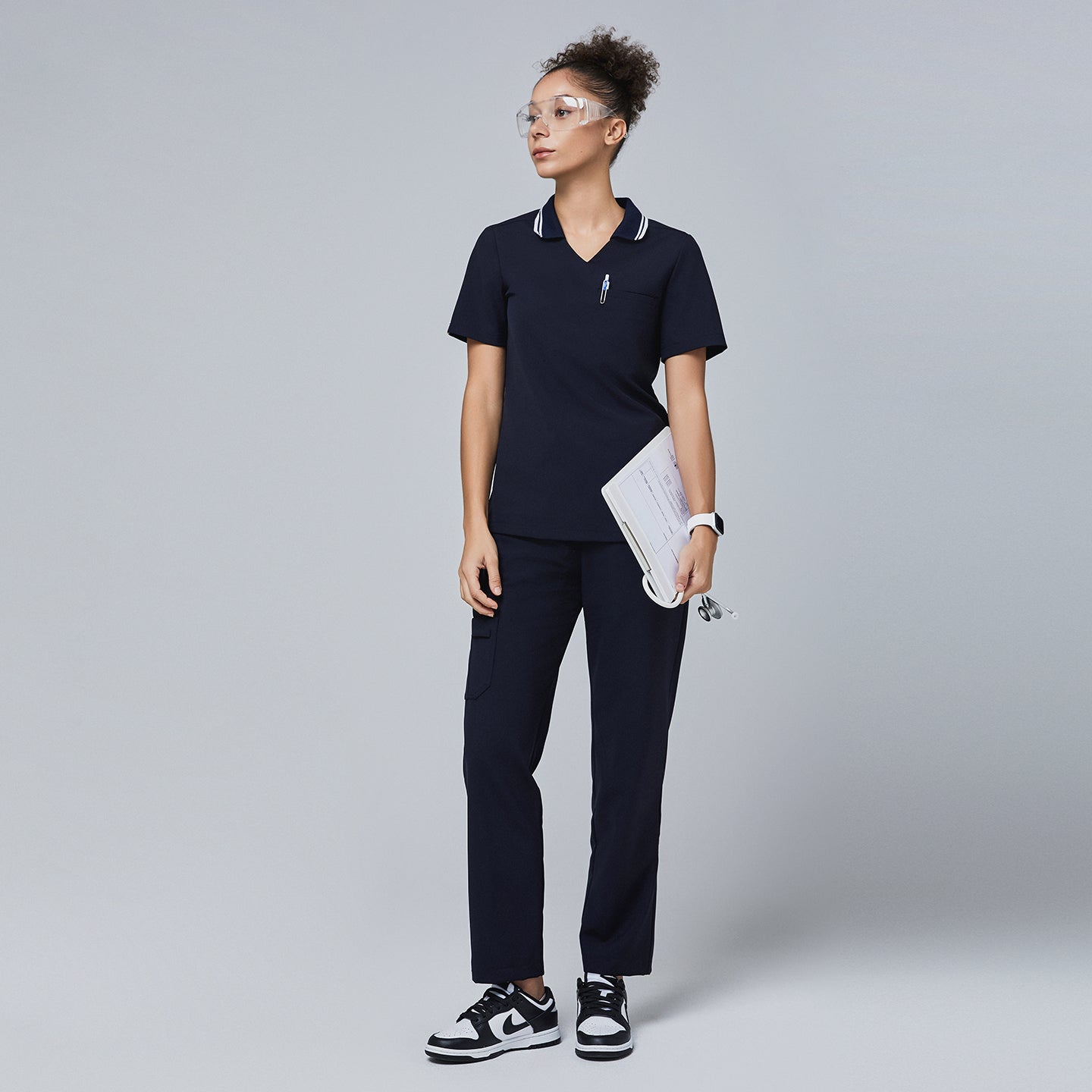 Woman in a navy blue polo scrub top with white striped collar and matching pants, holding a clipboard and stethoscope,Midnight Navy