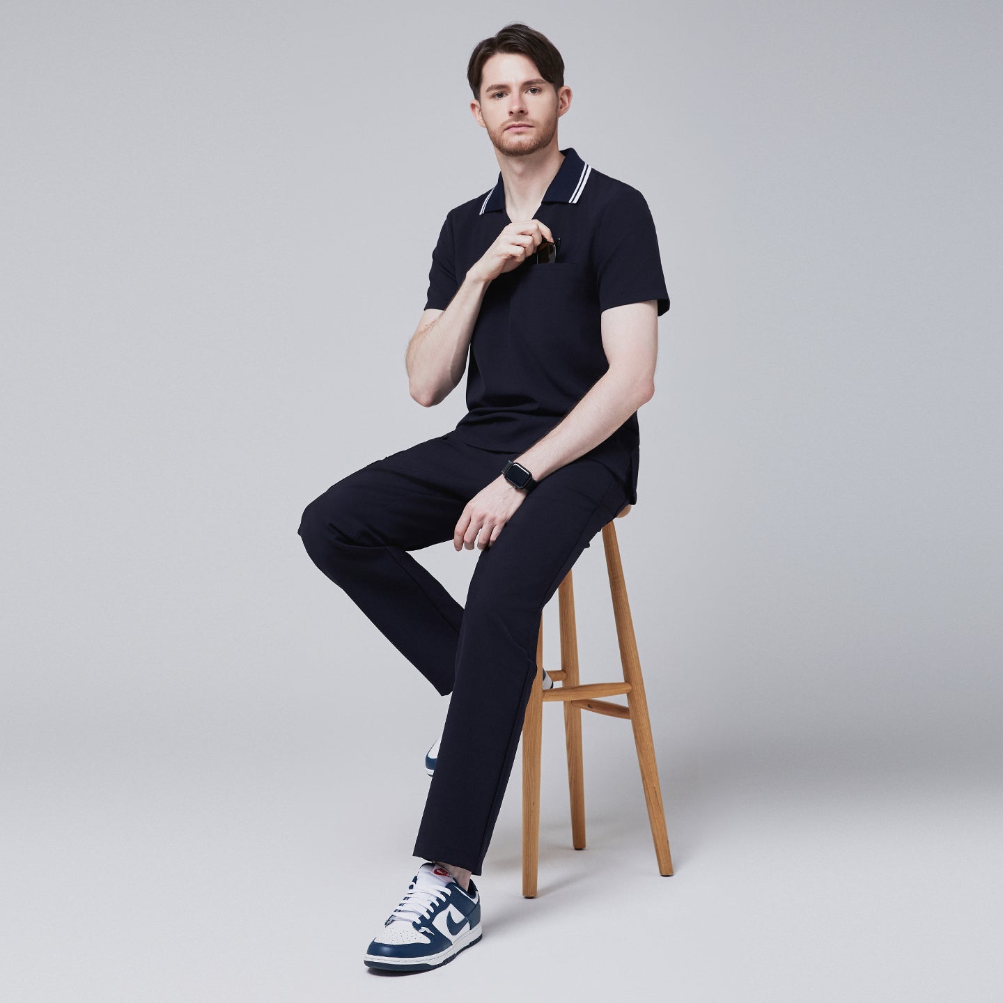 Male model sitting on a wooden stool wearing a midnight navy performance polo scrub top and matching pants, holding sunglasses,Midnight Navy