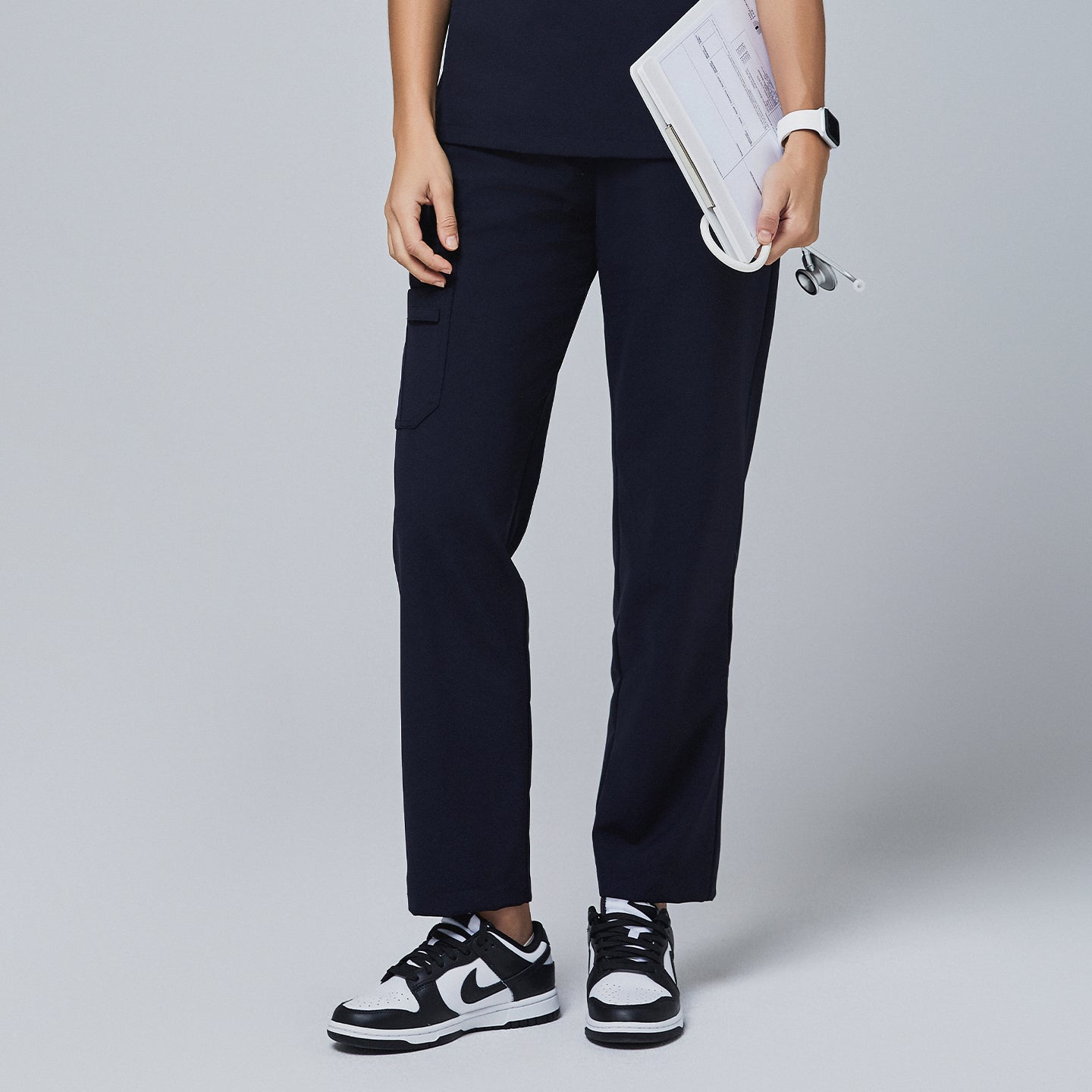 Navy utility scrub pants featuring a side pocket, worn with a navy top and paired with black-and-white sneakers, holding a clipboard and stethoscope,Midnight Navy