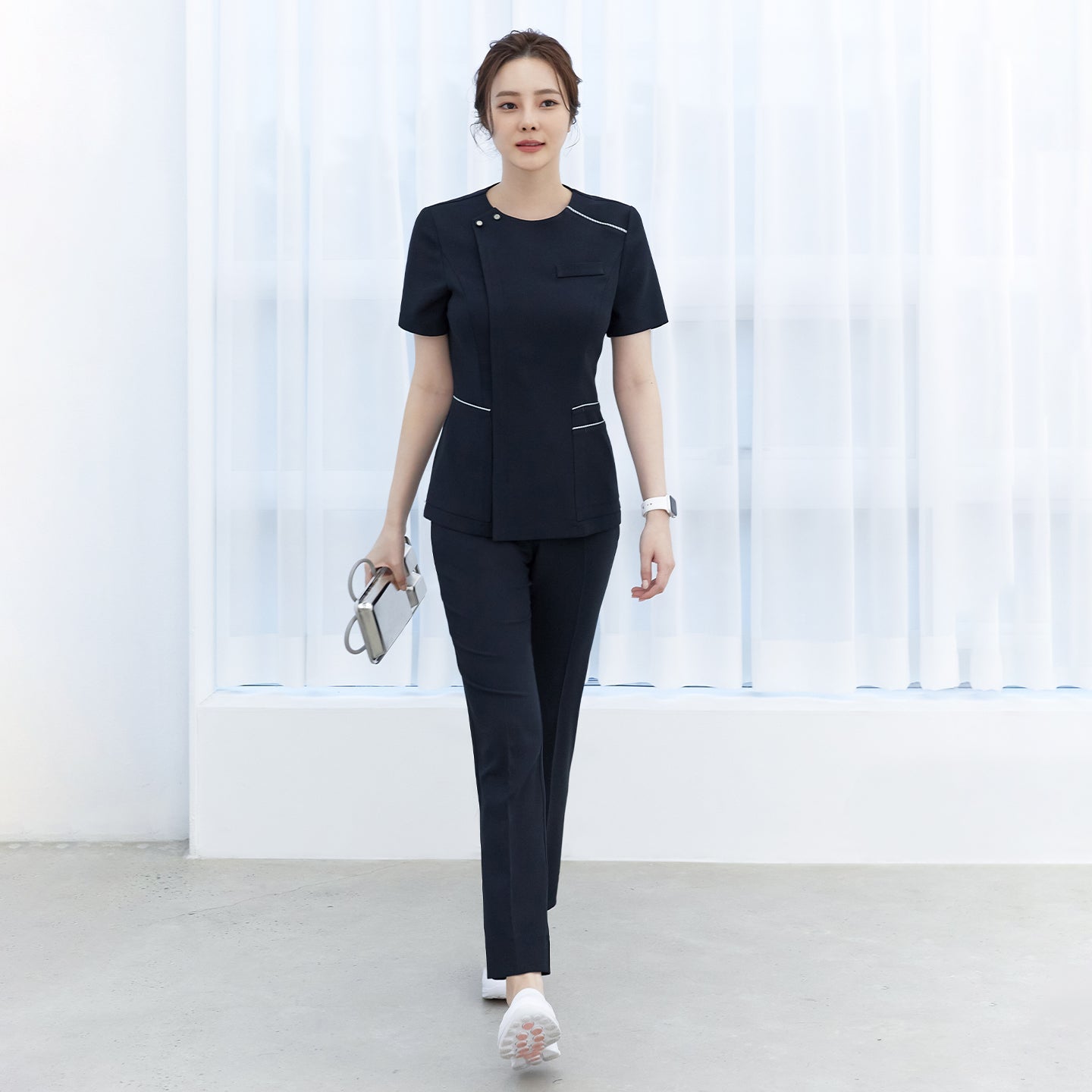 Woman walking in navy side-banding scrub pants with a matching navy top featuring white piping details and short sleeves, showing the full front view,Mir Navy