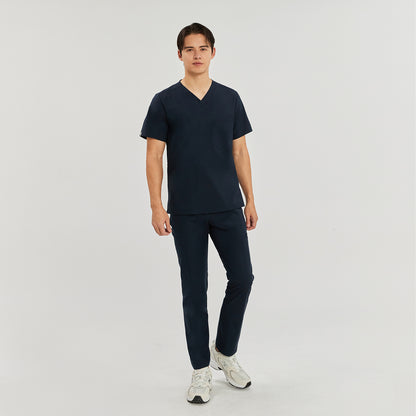Male model wearing a navy blue scrub top with a V-neck and a single chest pocket, paired with matching navy blue pants and white sneakers, standing against a plain background, Navy