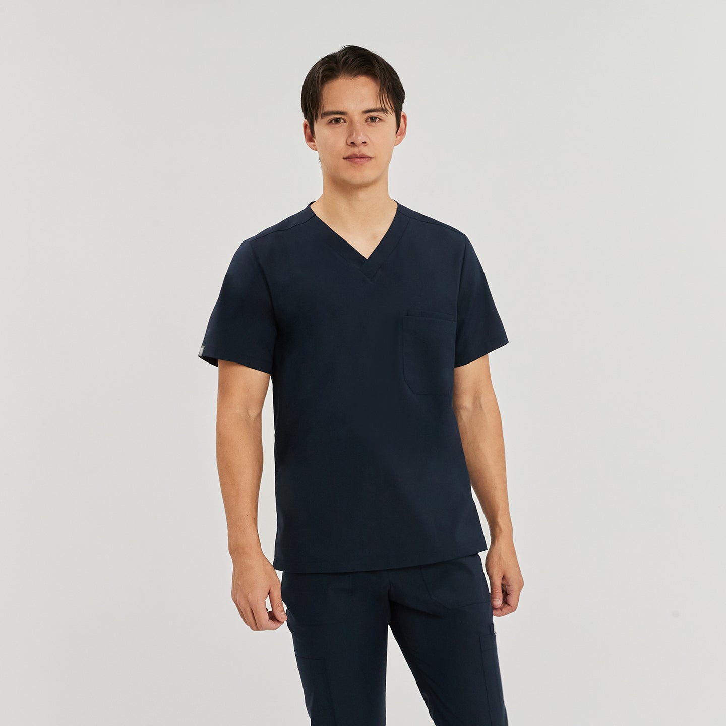 Male model wearing a navy blue scrub top with a V-neck and a single chest pocket, standing against a plain background, Navy