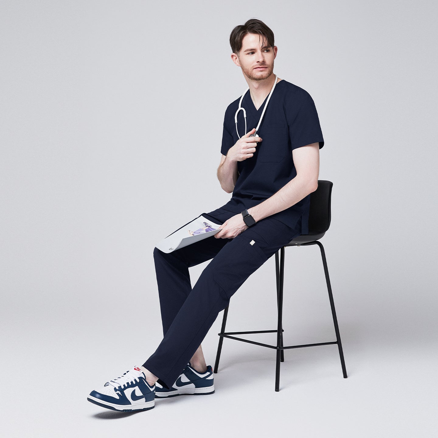 man seated, wearing Zenir navy 8-pocket scrub pants, matching top, stethoscope, and blue sneakers, holding a clipboard,Navy