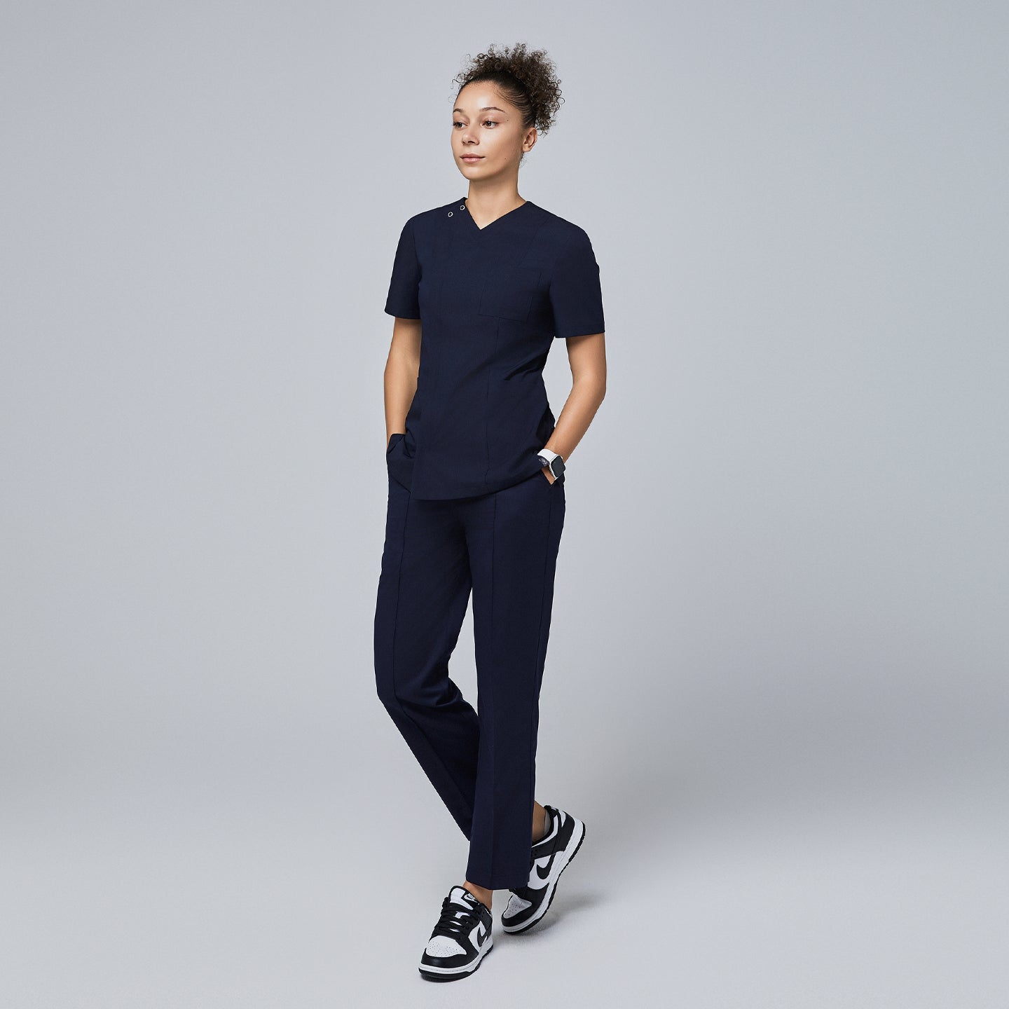 Female model wearing a navy V-neck scrub top with a front zipper, chest pocket, and short sleeves, paired with matching navy scrub pants and sneakers, standing with hands in pockets,Eco Navy