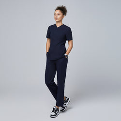 Image of Female model wearing a navy V-neck scrub top with a front zipper, chest pocket, and short sleeves, paired with matching navy scrub pants and sneakers, standing with hands in pockets,Eco Navy