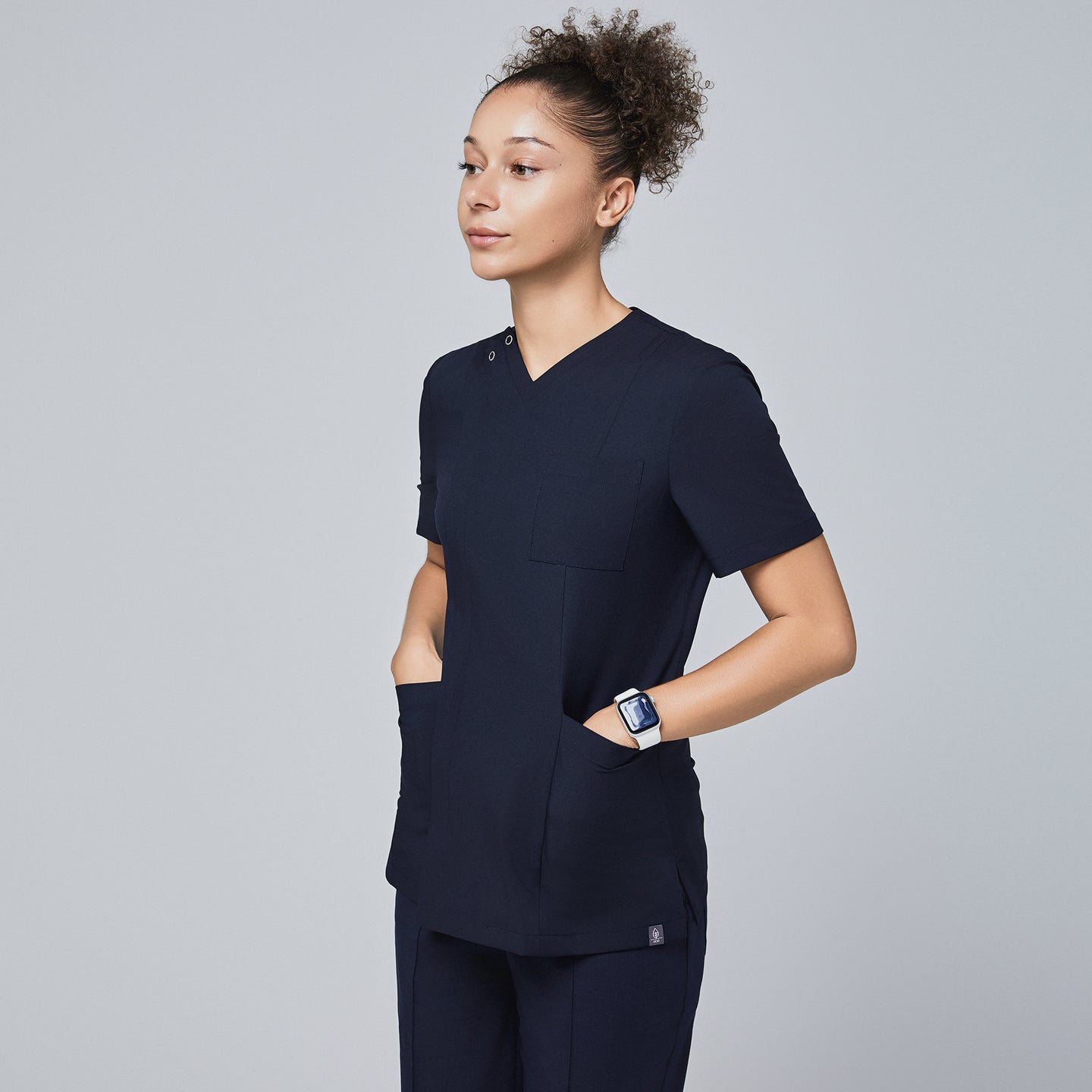 Female model wearing a navy V-neck scrub top with a front zipper, chest pocket, and two spacious front pockets, shown in a side profile with hands in pockets,Eco Navy