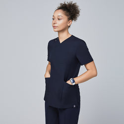 Image of Female model wearing a navy V-neck scrub top with a front zipper, chest pocket, and two spacious front pockets, shown in a side profile with hands in pockets,Eco Navy