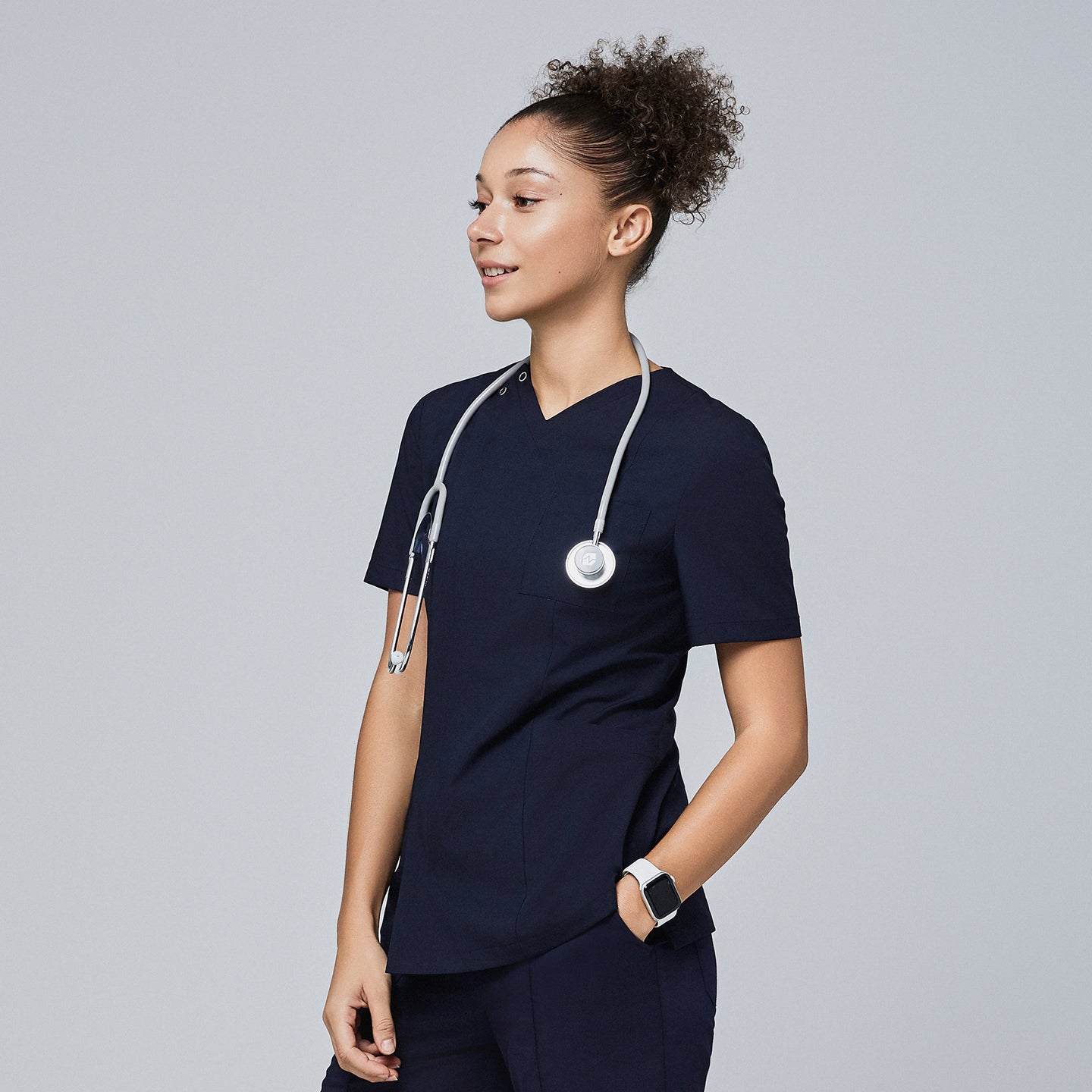 Female model wearing a navy V-neck scrub top with a front zipper, chest pocket, and short sleeves, accessorized with a stethoscope around her neck, smiling and looking to the side,Eco Navy