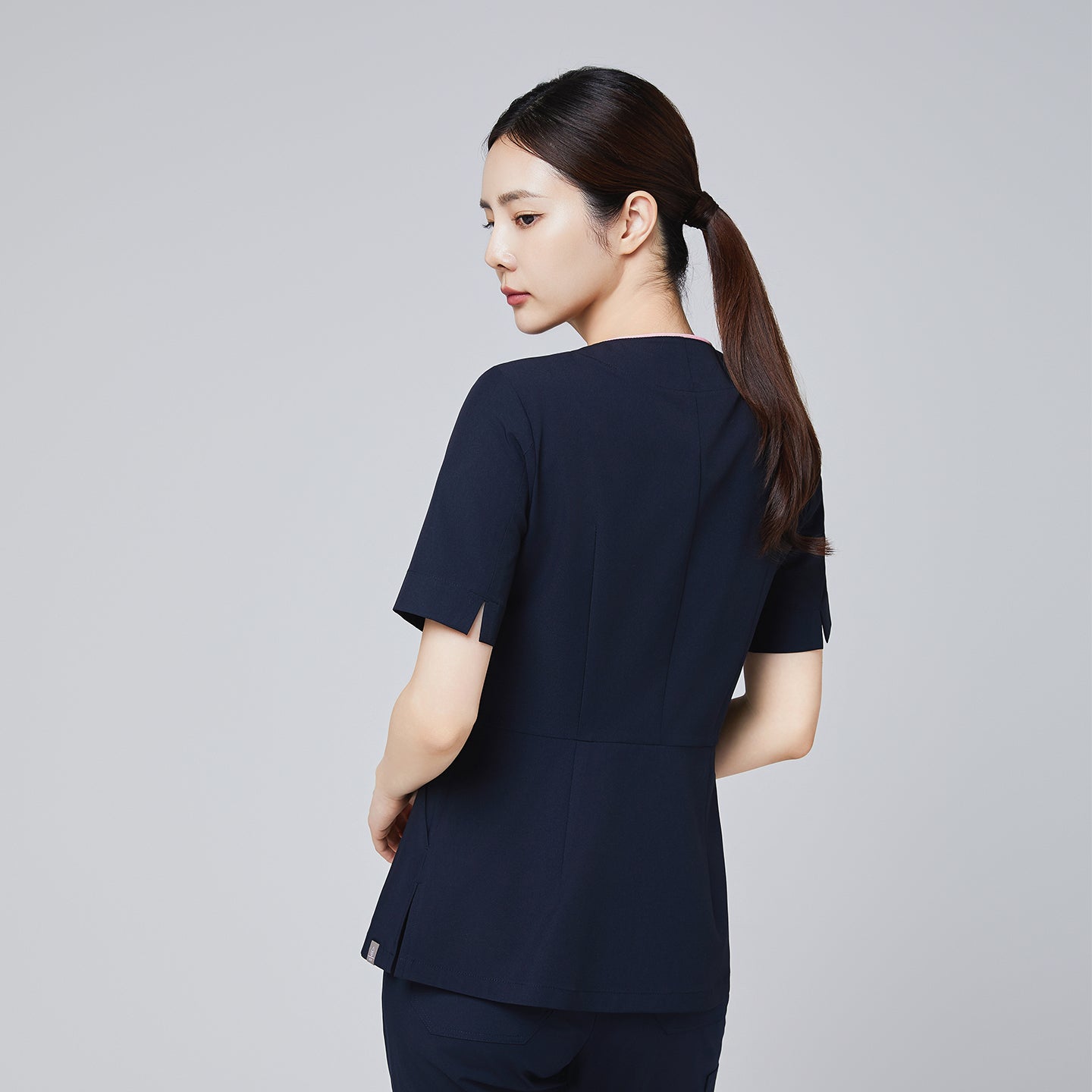 Back view of a navy scrub top with short sleeves and tailored seams, modeled by a woman in a professional pose.Navy