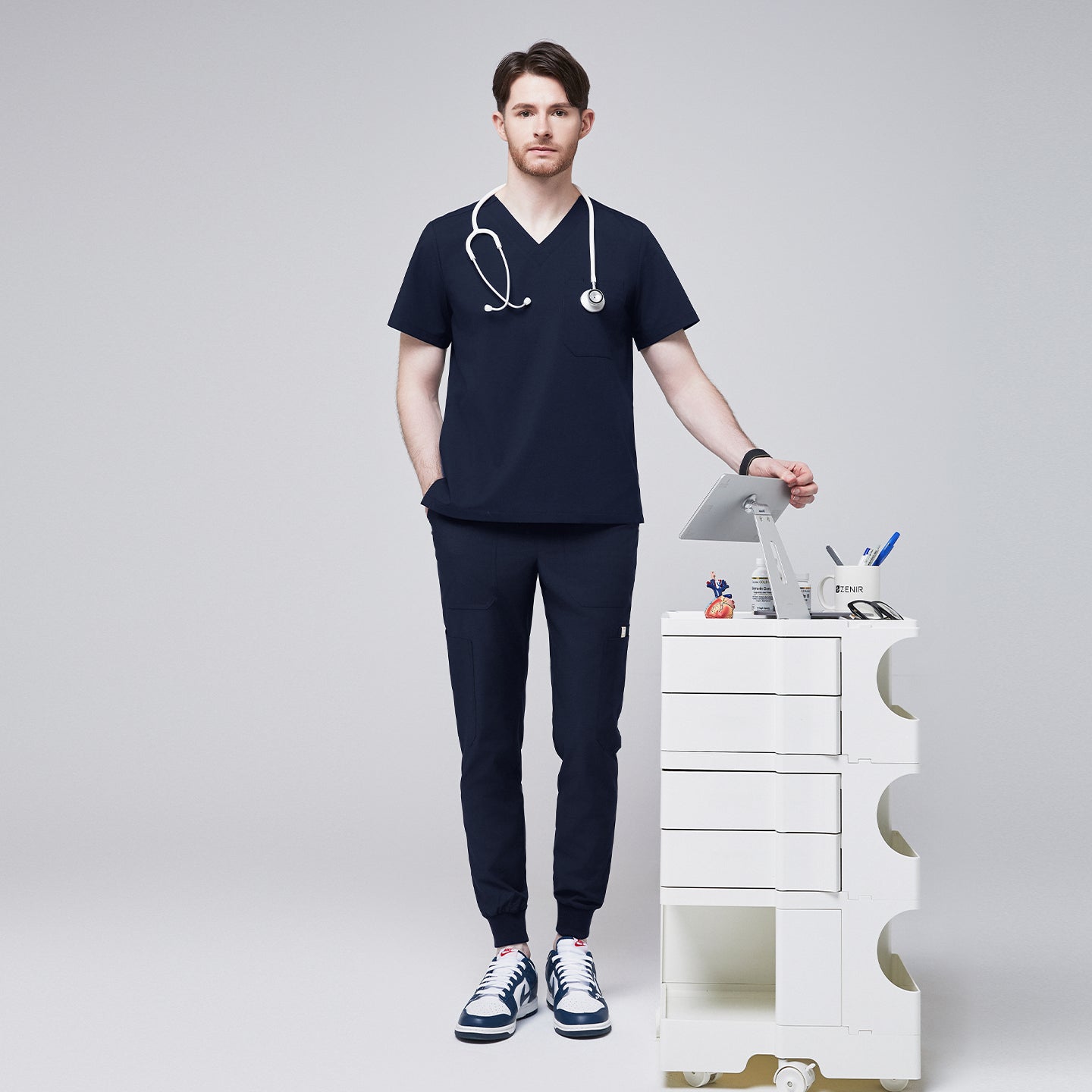 Male healthcare professional wearing a navy V-neck, three-pocket scrub top and matching jogger pants with stethoscope around neck, standing beside a medical cart with supplies,Navy